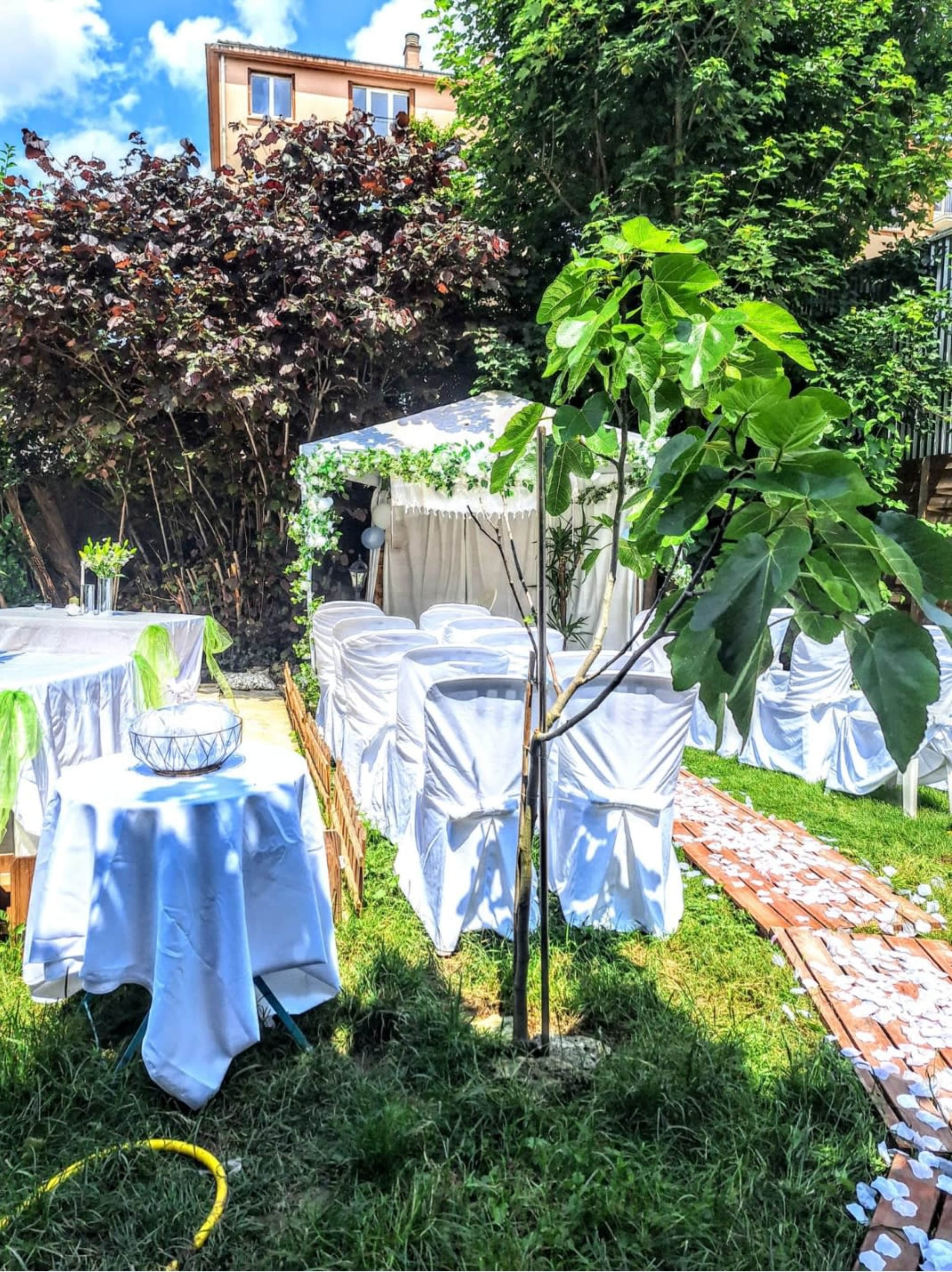 A garden setting features white table covers, a canopied area, and greenery, with several chairs arranged around tables.