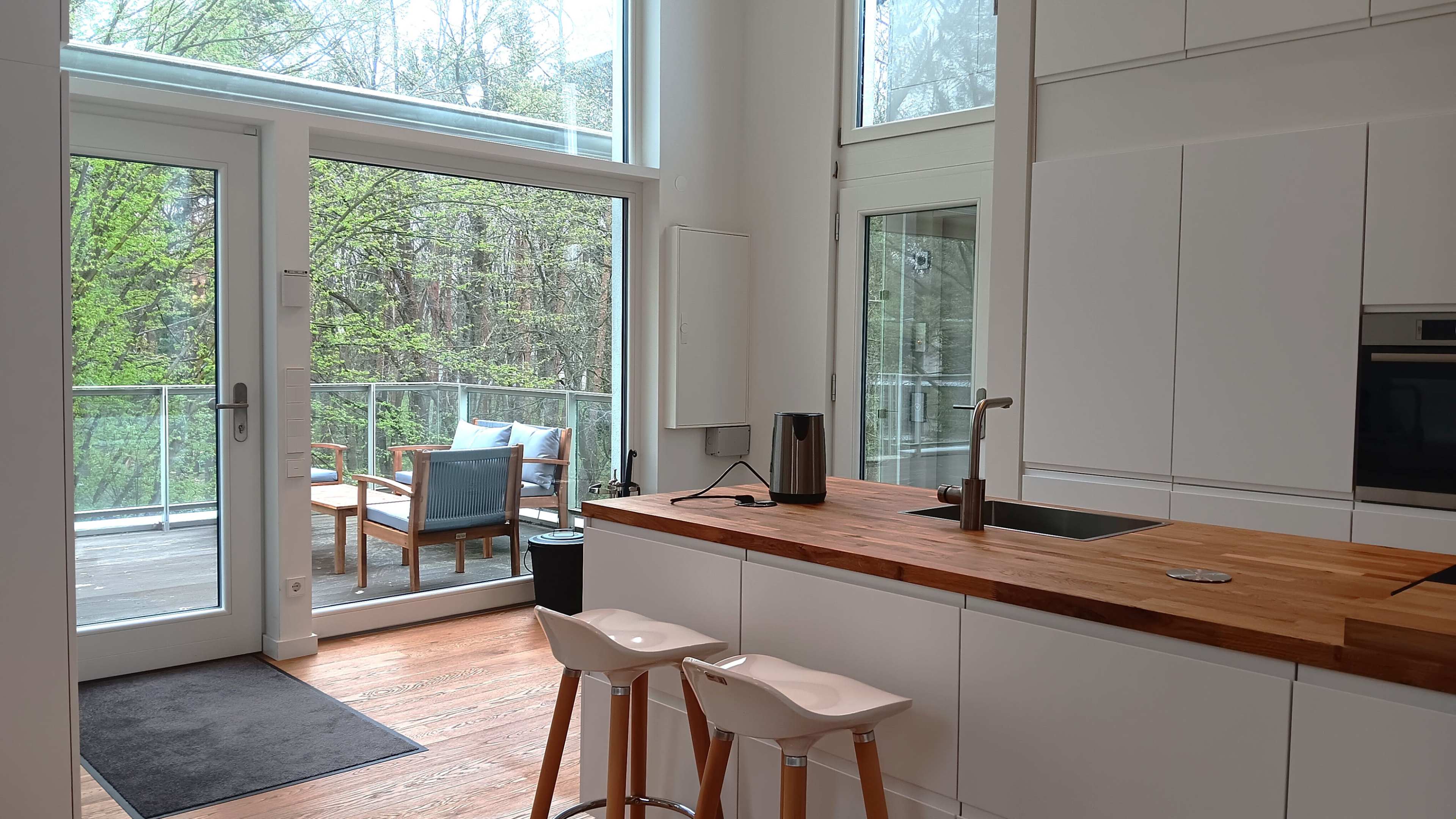 A modern kitchen features a wooden countertop and two high stools, with large glass doors leading to an outdoor seating area surrounded by greenery.