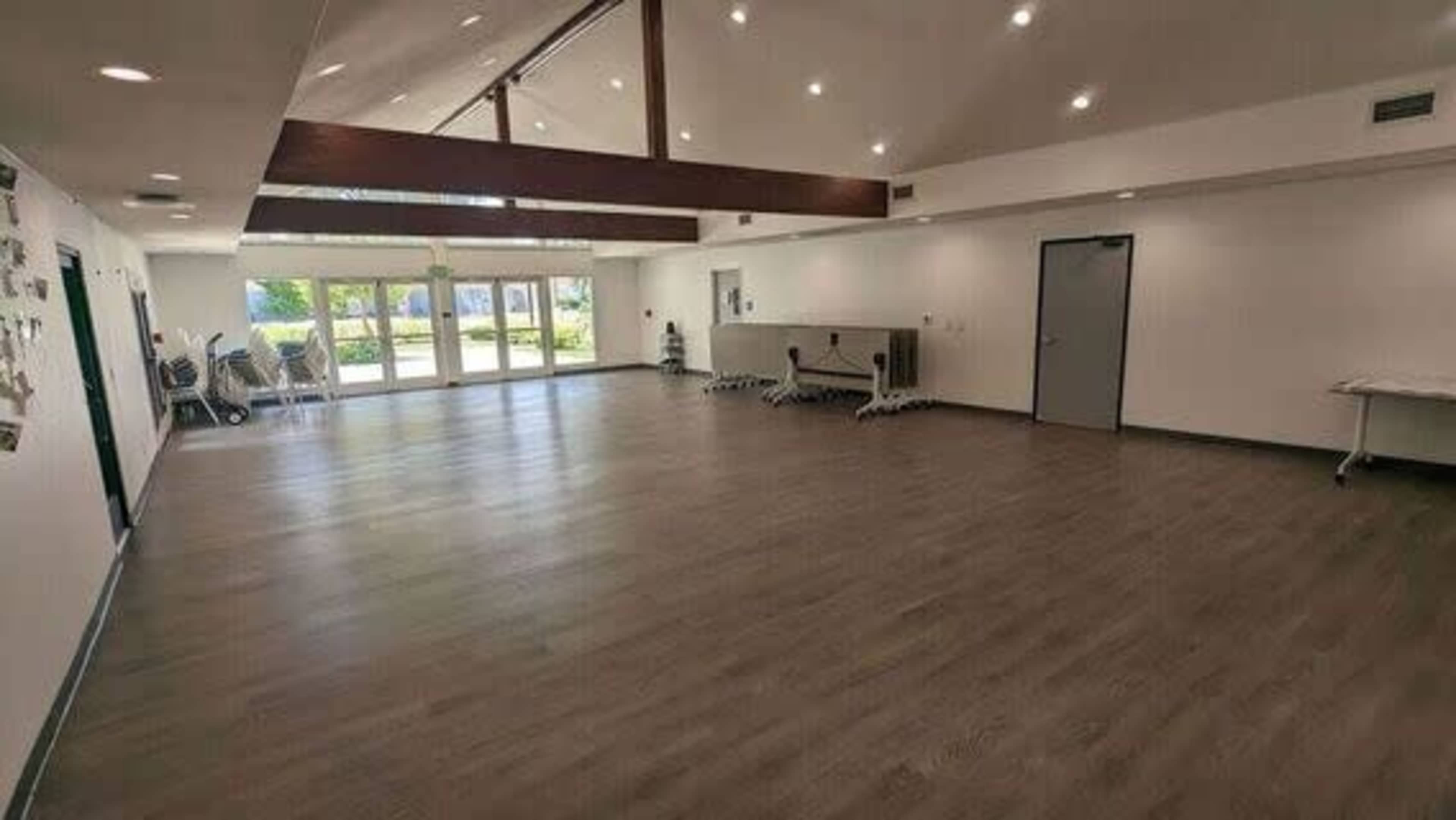 A spacious, brightly lit room with wooden flooring and large windows, featuring exercise equipment along one wall.