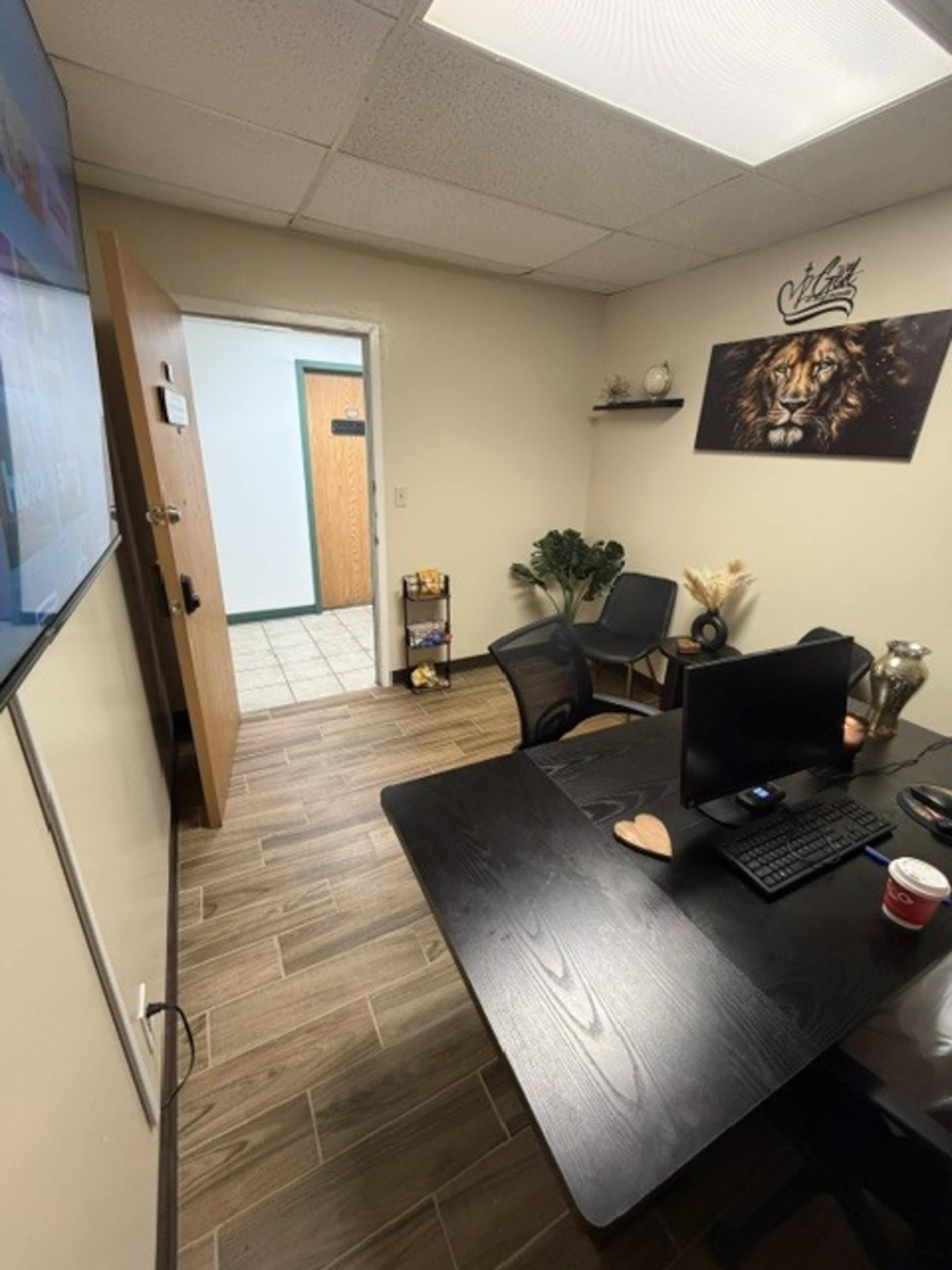 The image shows a small office space with a black desk, a computer, and two chairs, along with a door leading to a corridor and a wall featuring a lion-themed decoration.