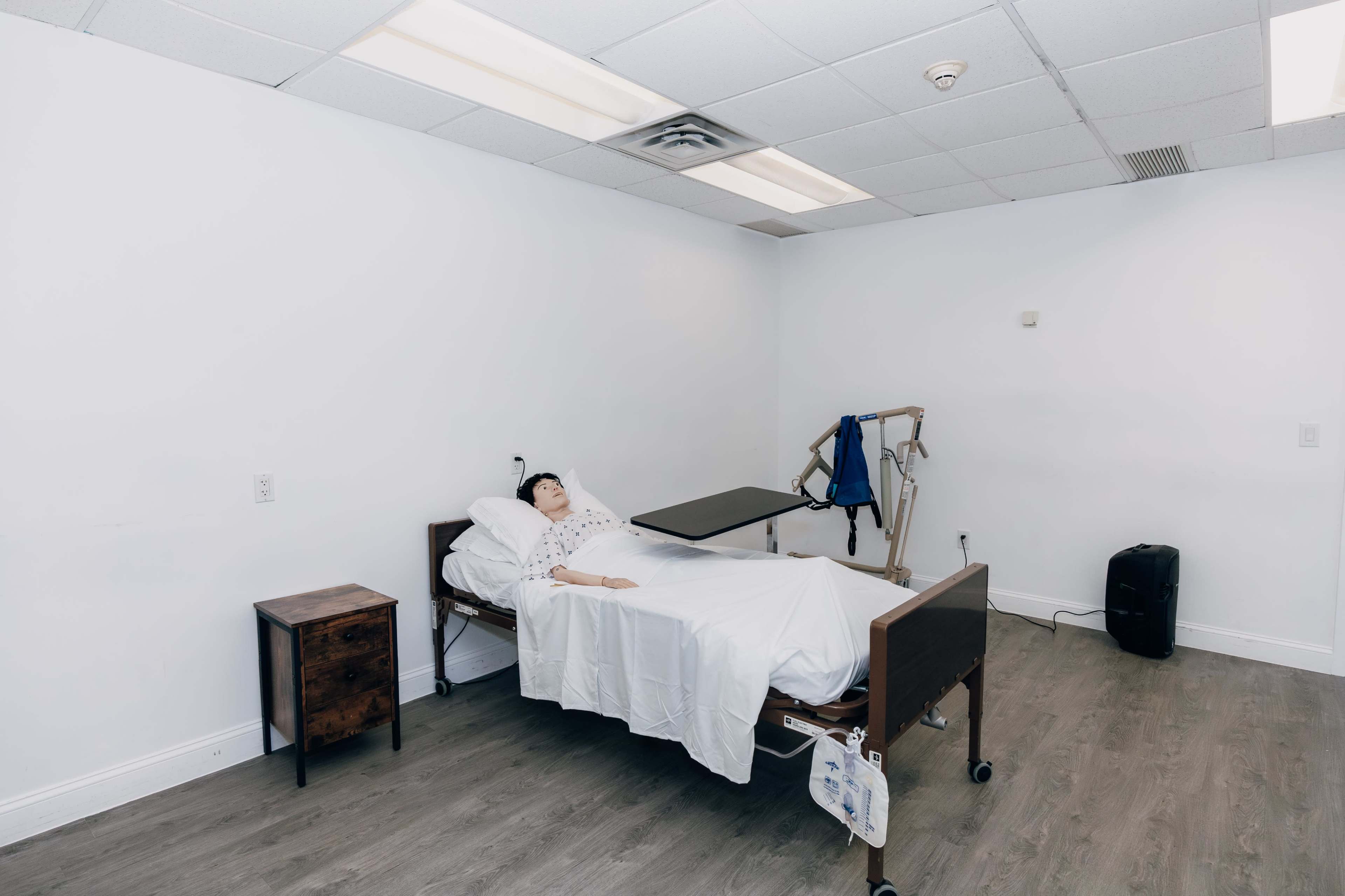 The image shows a hospital room with a bed and a mannequin lying under a white blanket, accompanied by a bedside table and a mobility aid.
