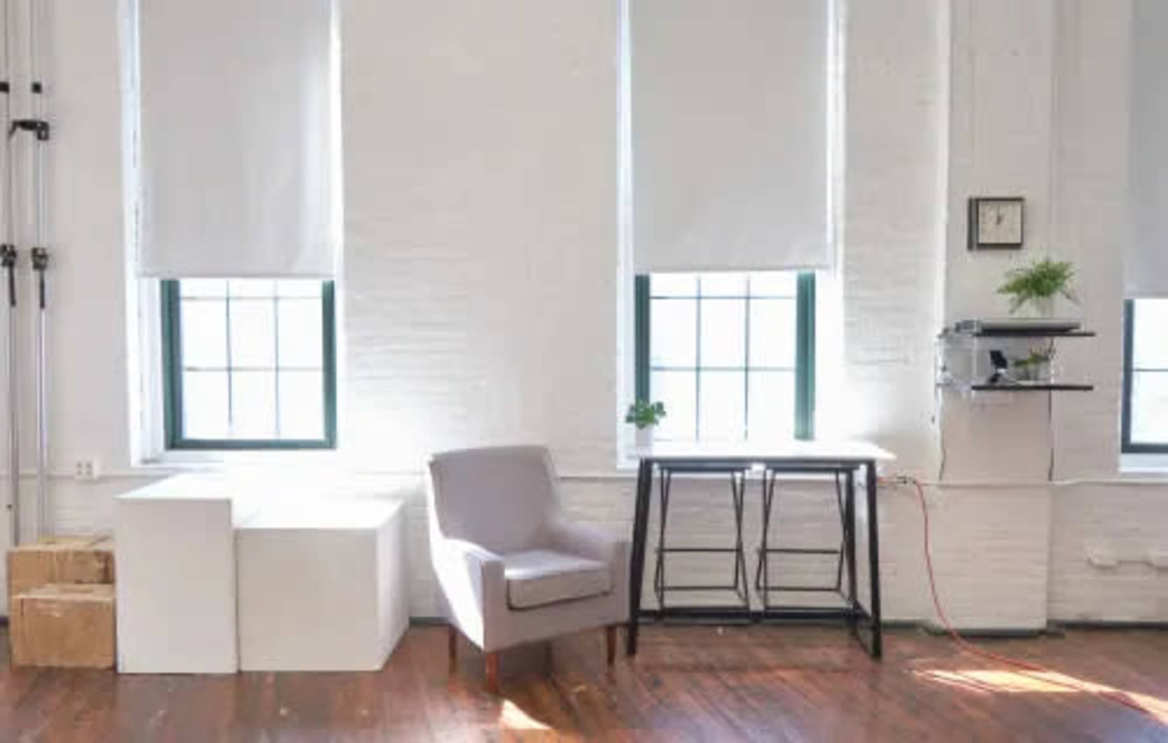 The scene features a minimalist interior with two windows, a gray armchair, a small black table, and a white wall.