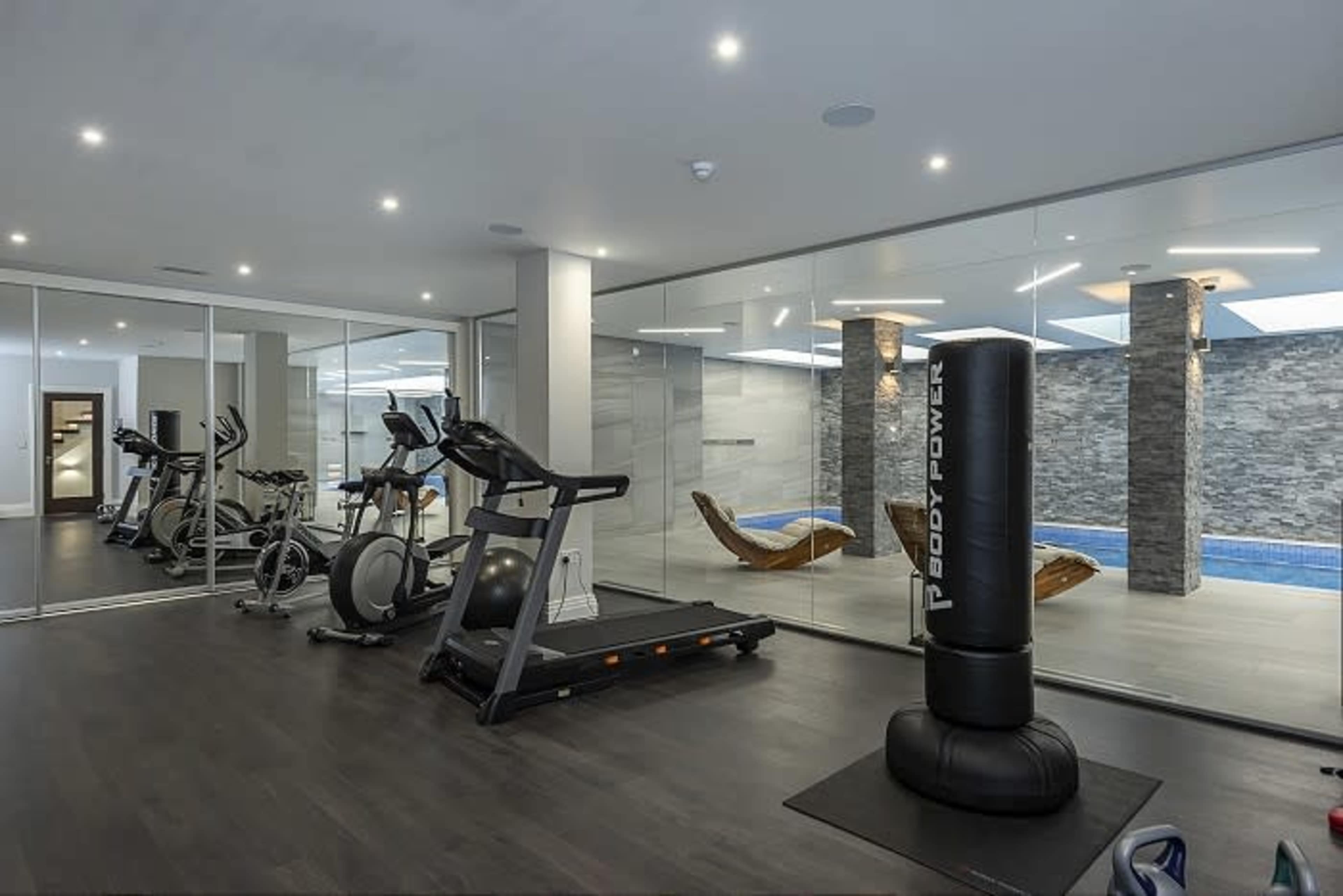 The image shows a modern gym area with exercise equipment and a glass wall overlooking a pool.