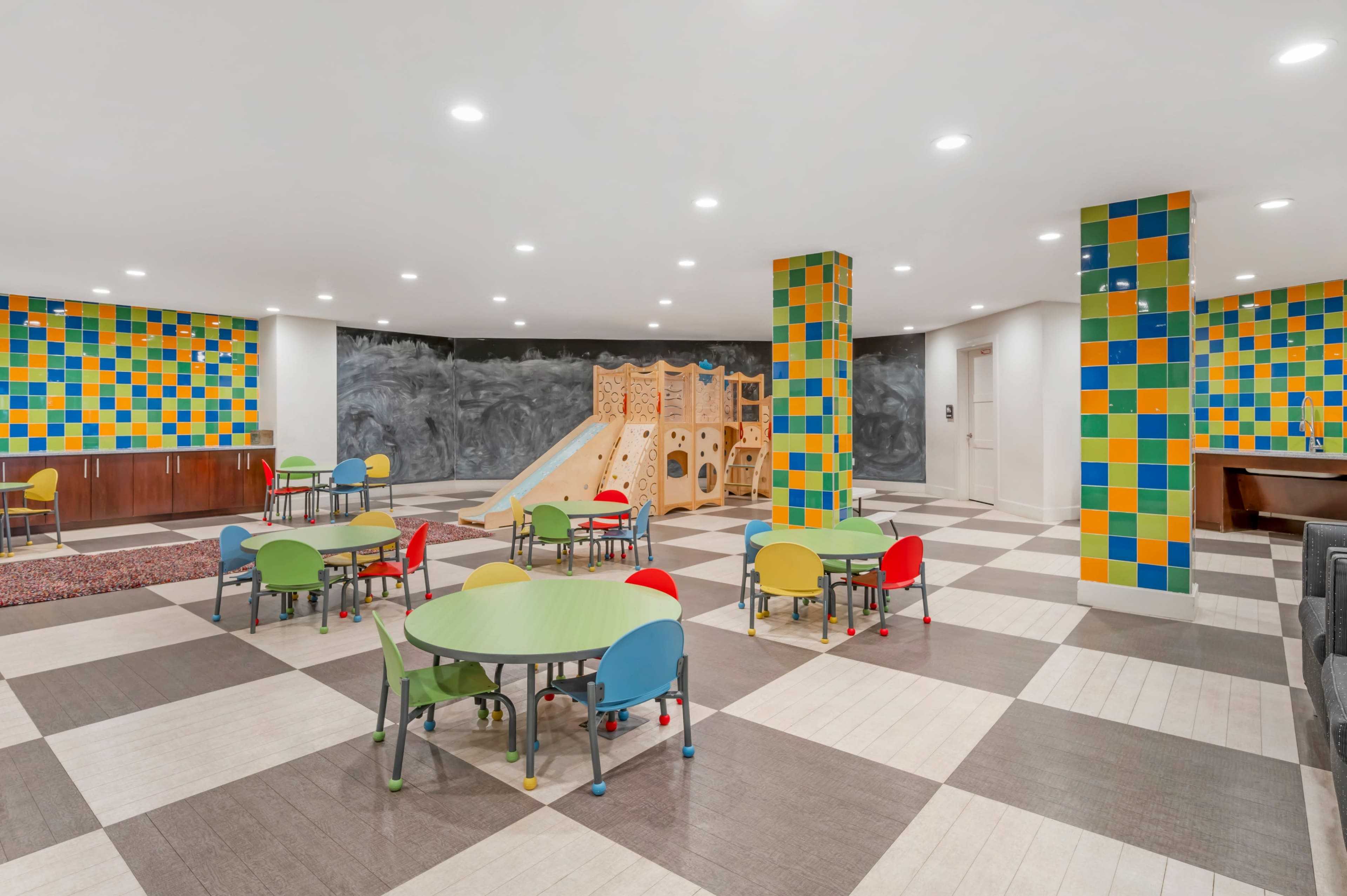 The image shows a colorful playroom featuring a climbing structure, several small tables with chairs, and patterned walls.