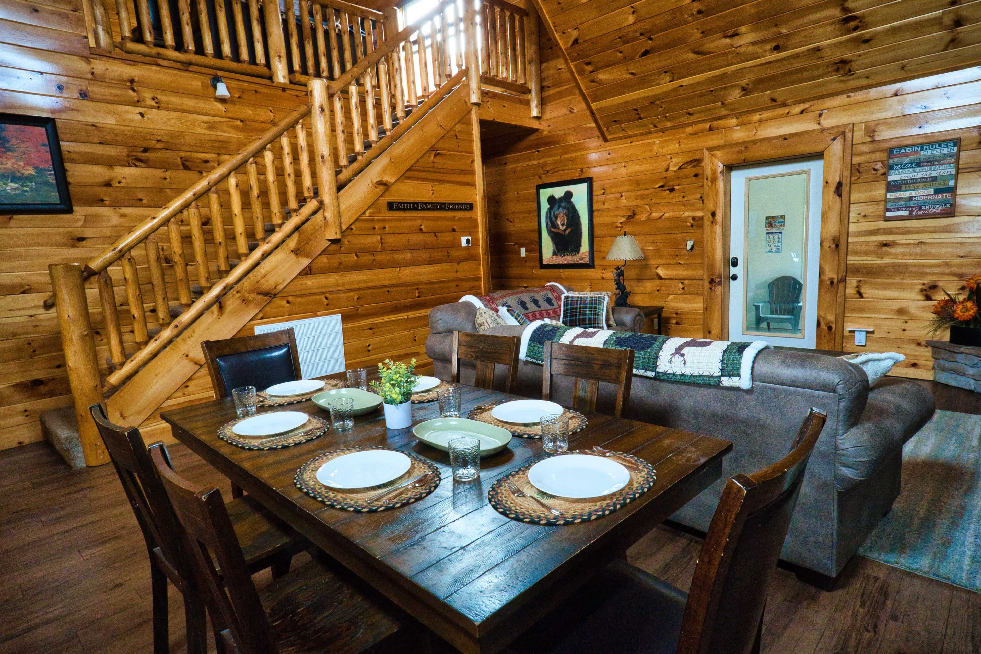 The image shows a wooden cabin interior with a dining table set for a meal, a sofa, and a staircase in the background.