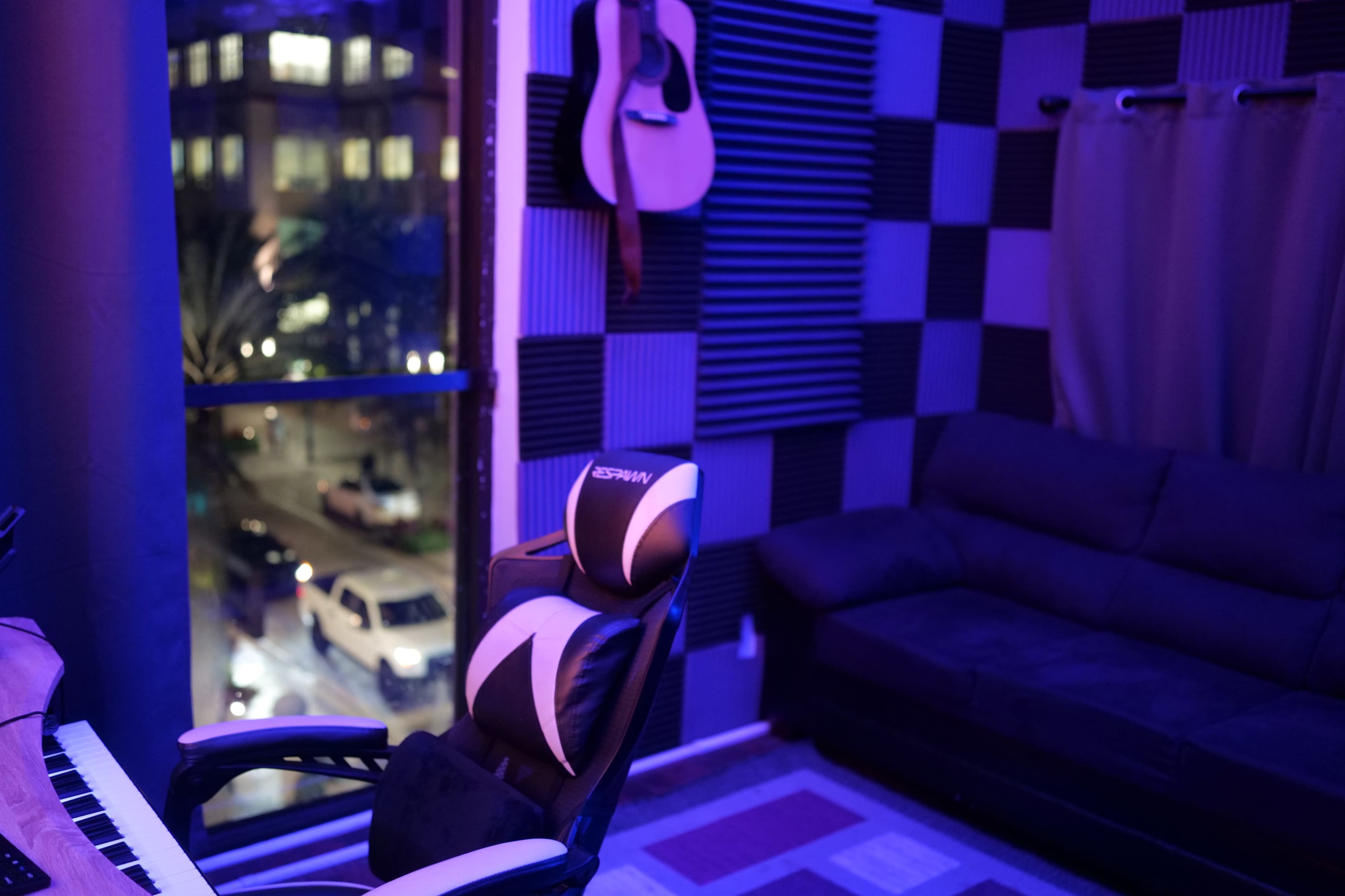 A dimly lit room with a keyboard, a gaming chair, a guitar hanging on the wall, and a view of a street outside through a large window.
