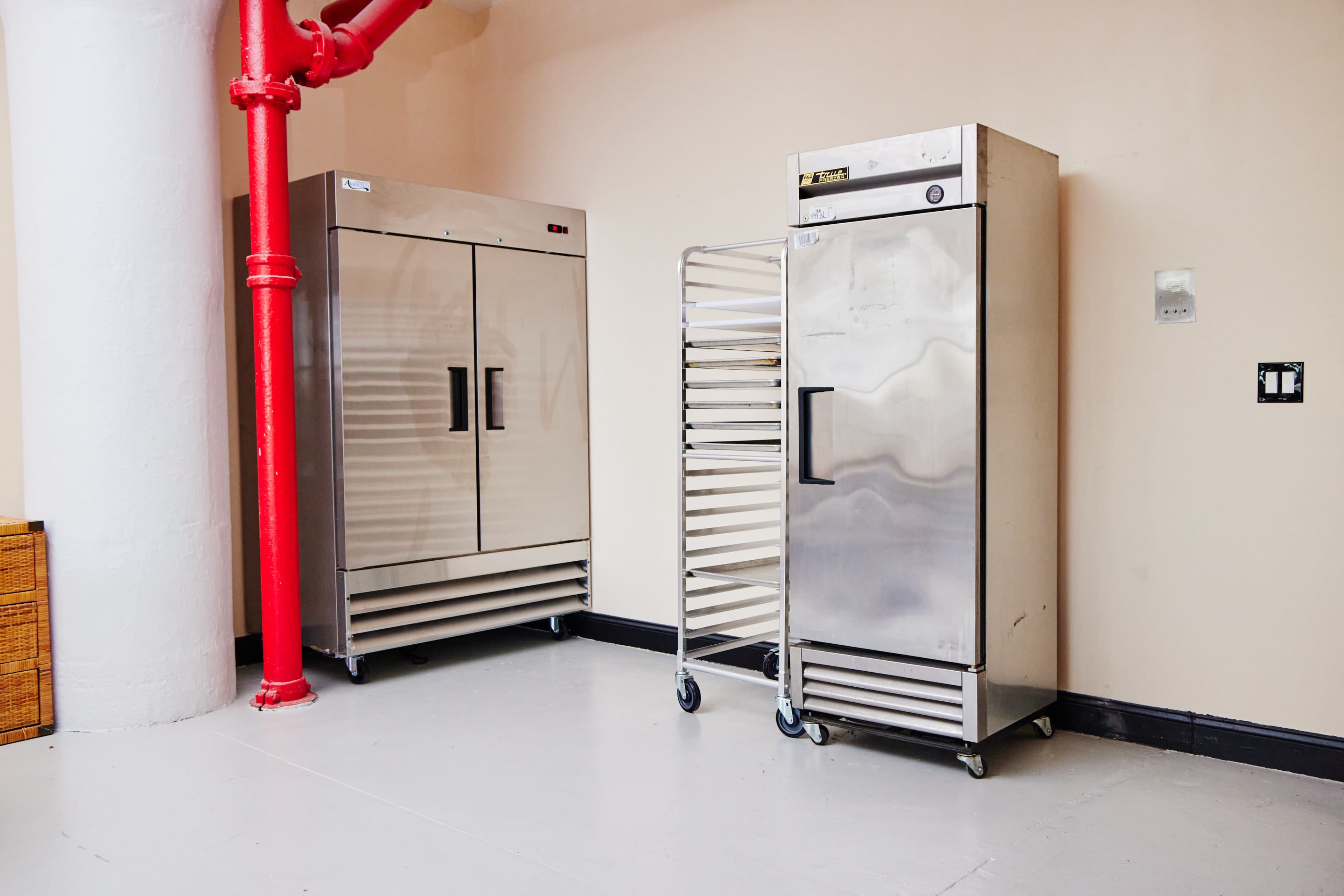 The image shows two stainless steel refrigerators and a metal rack on wheels in a room with a light-colored wall and a red pipe.