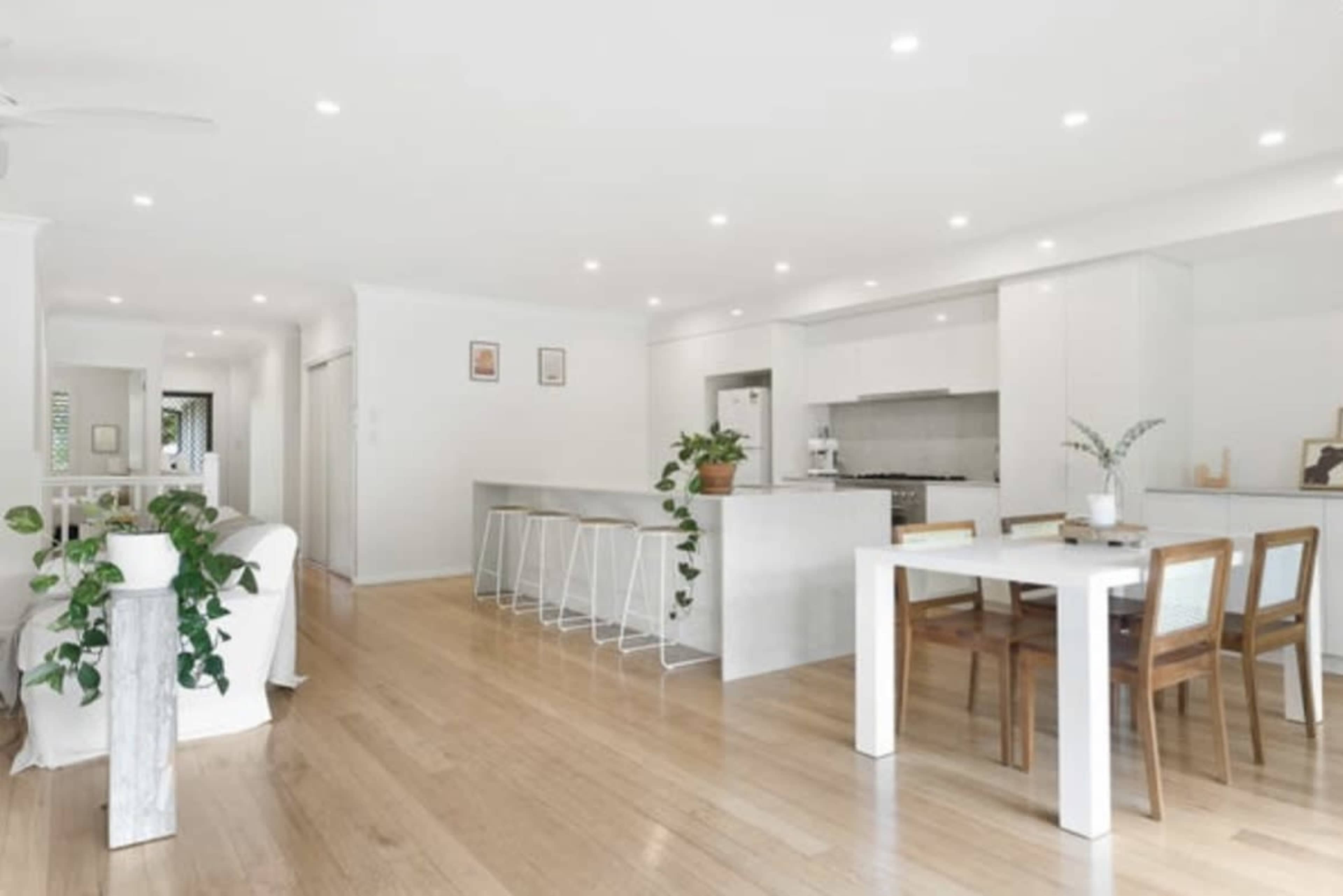 The image shows a modern open-plan living and dining area with a kitchen, featuring light wood flooring and minimalist furniture.