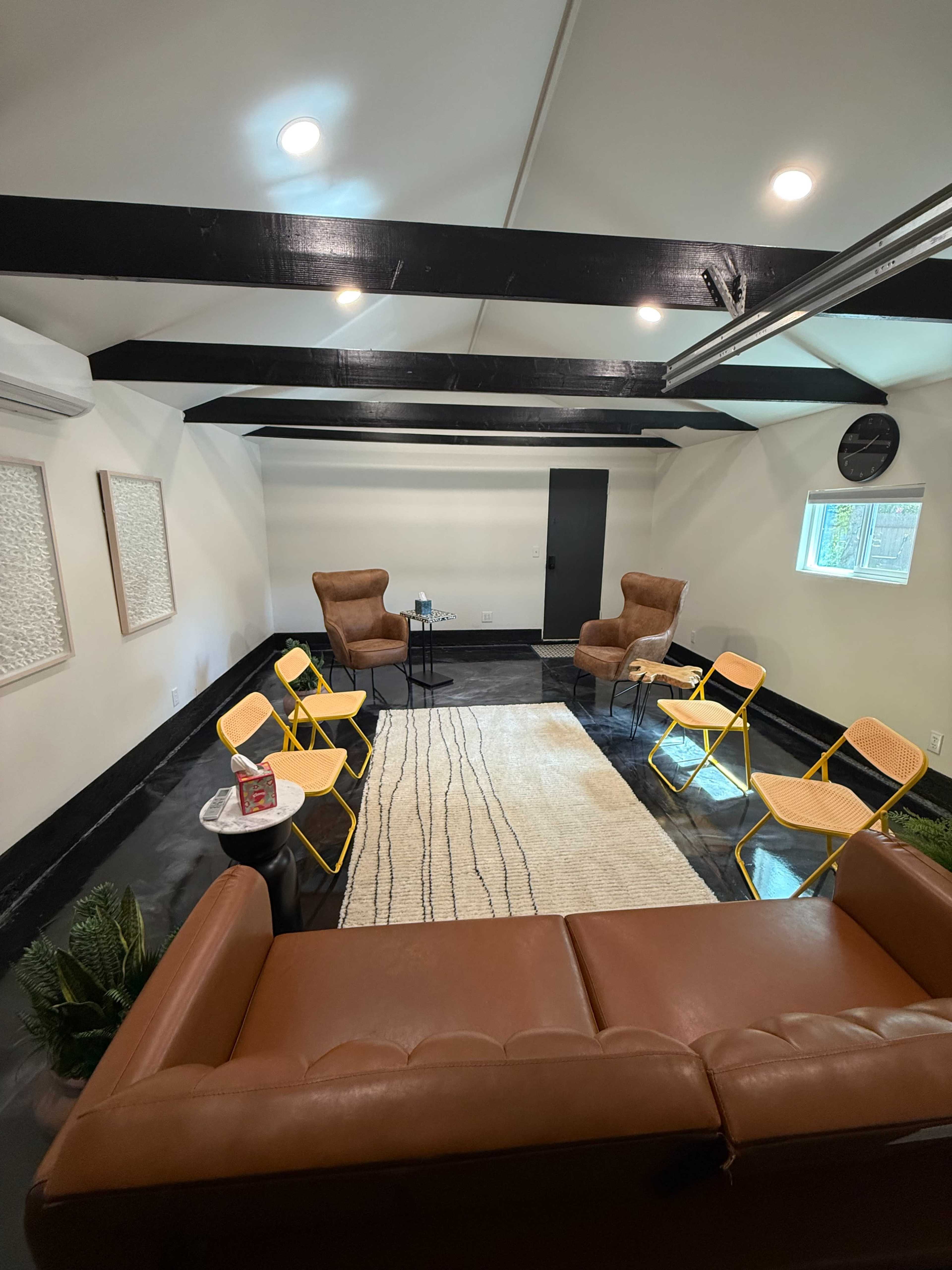The image shows a modern room with brown leather furniture, two armchairs, four yellow folding chairs, a small table, and a rug placed in the center under exposed beams.