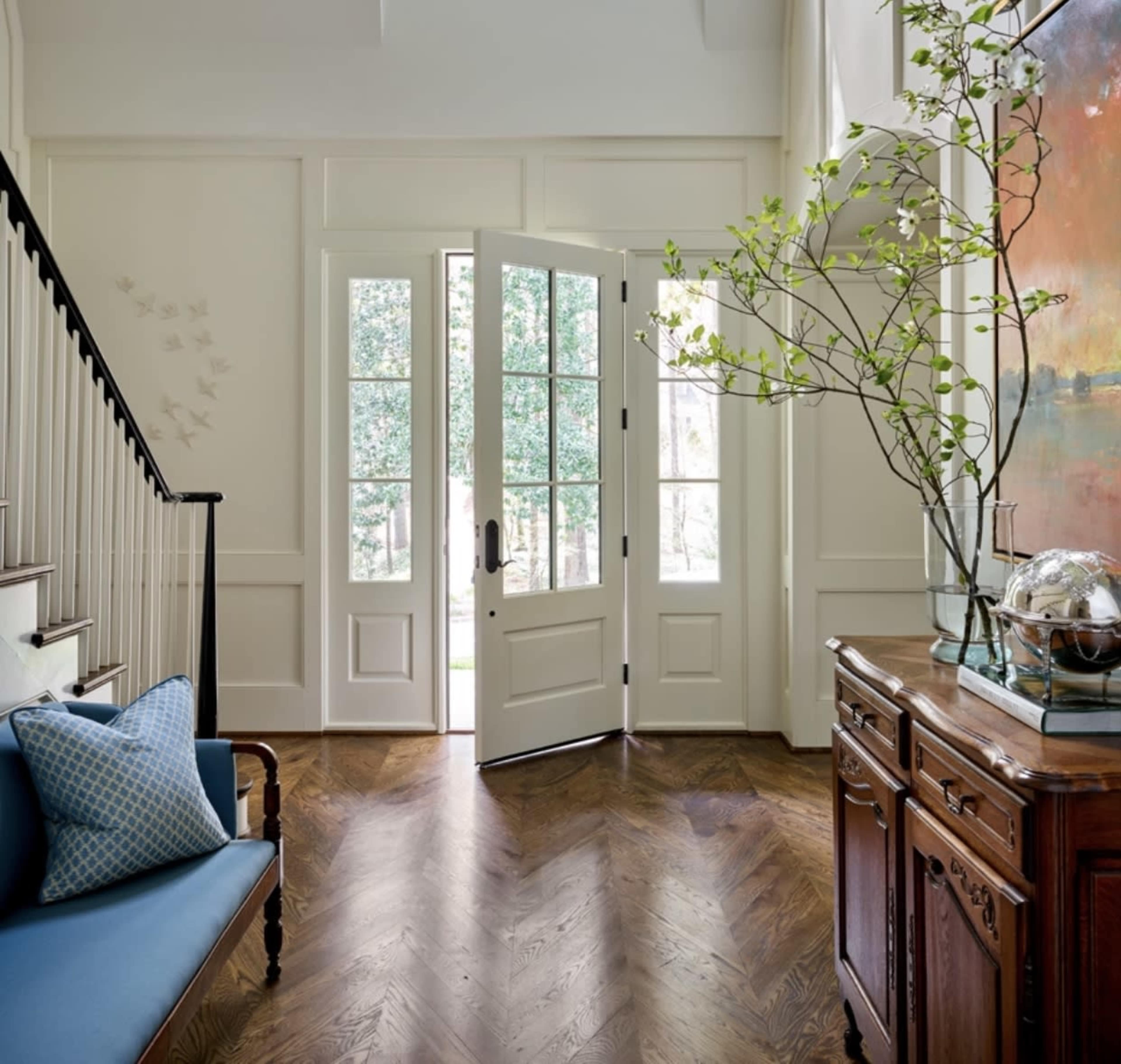 A spacious entryway features a double front door open to a view of the outdoors, with a blue bench and a wooden side table nearby.