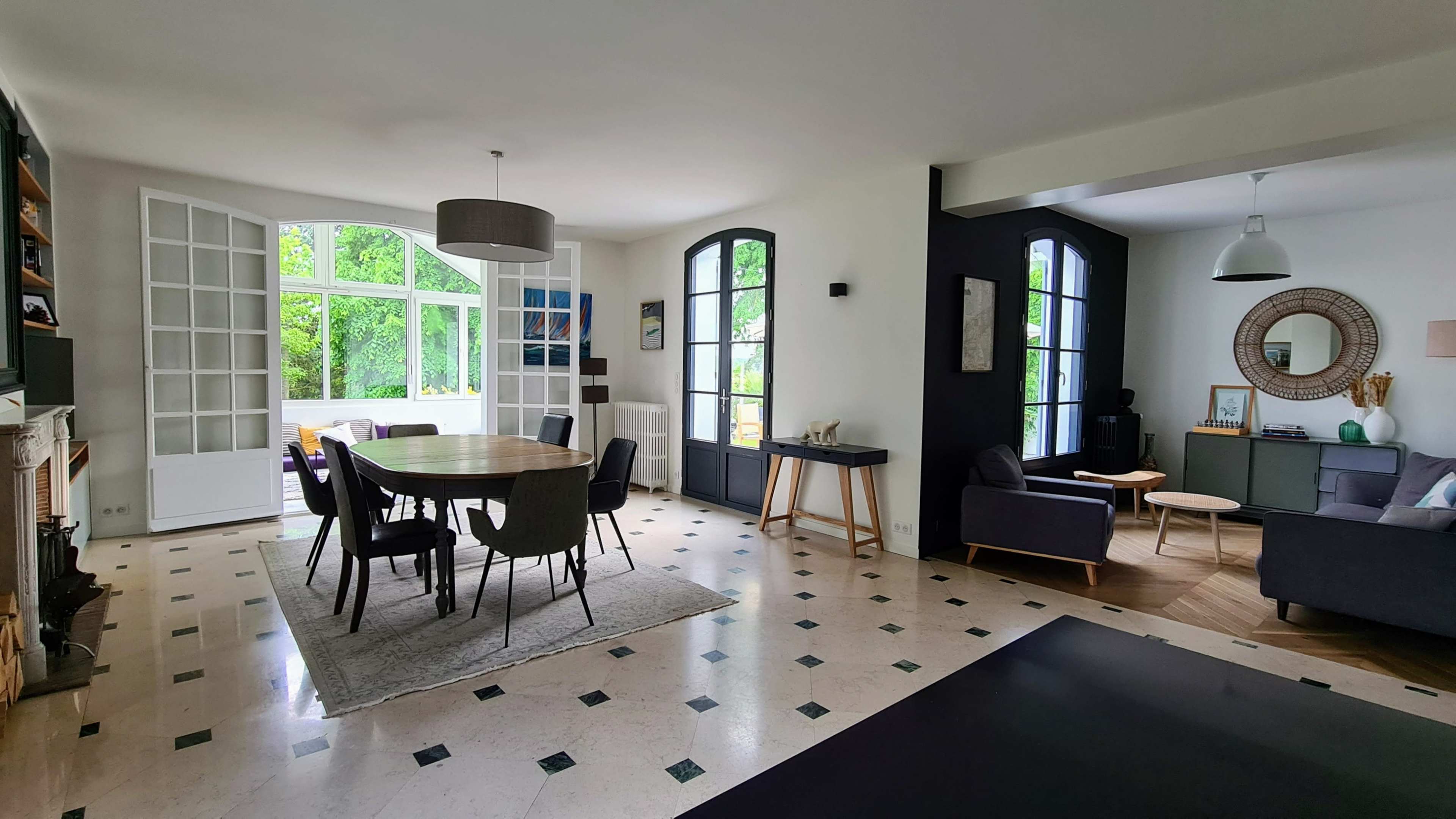 A modern, open dining and living area featuring a circular dining table surrounded by chairs, with large windows letting in natural light and a mix of furniture styles.