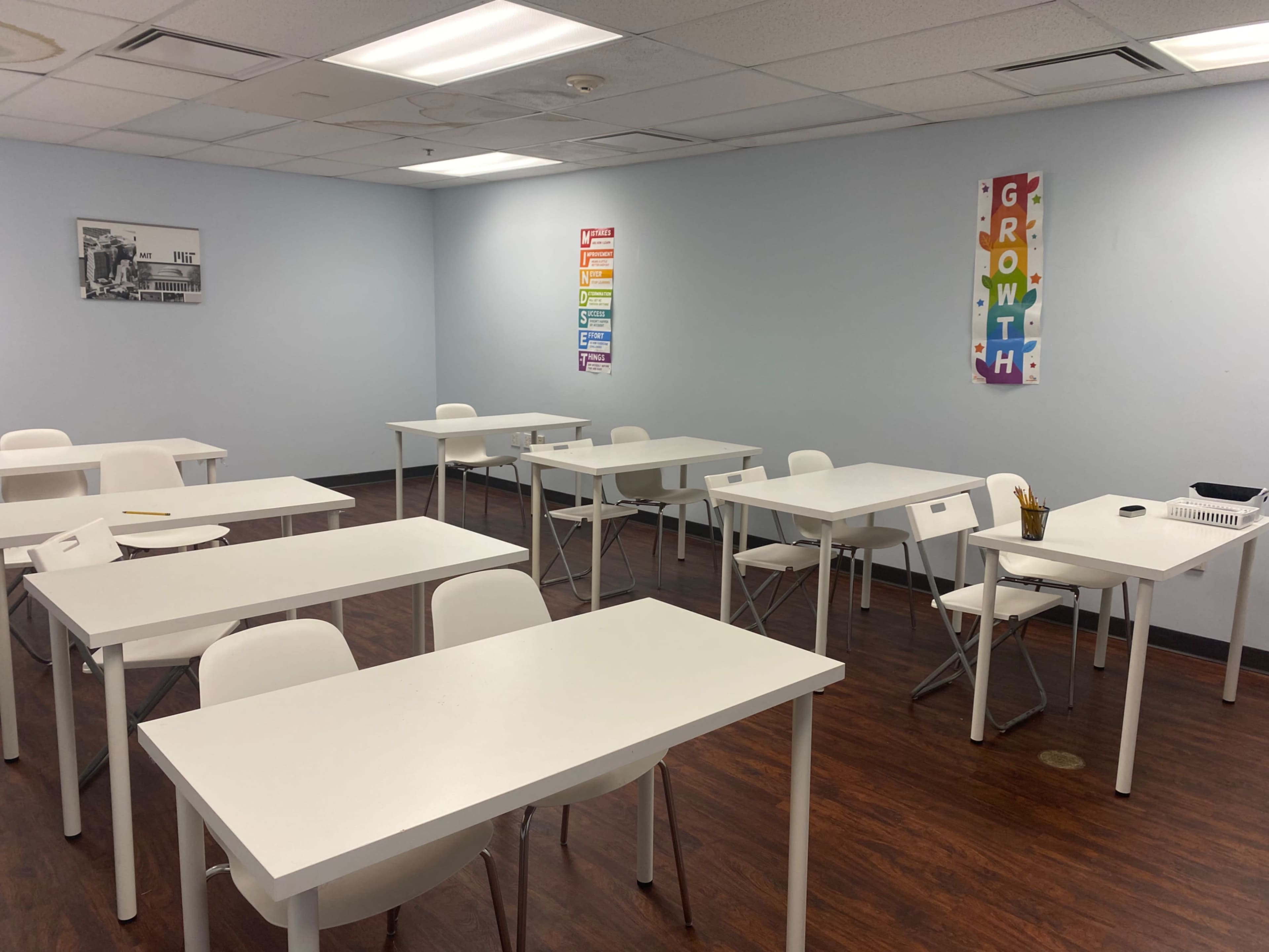 A classroom with white tables and chairs arranged in a sparse layout, featuring posters on the walls.