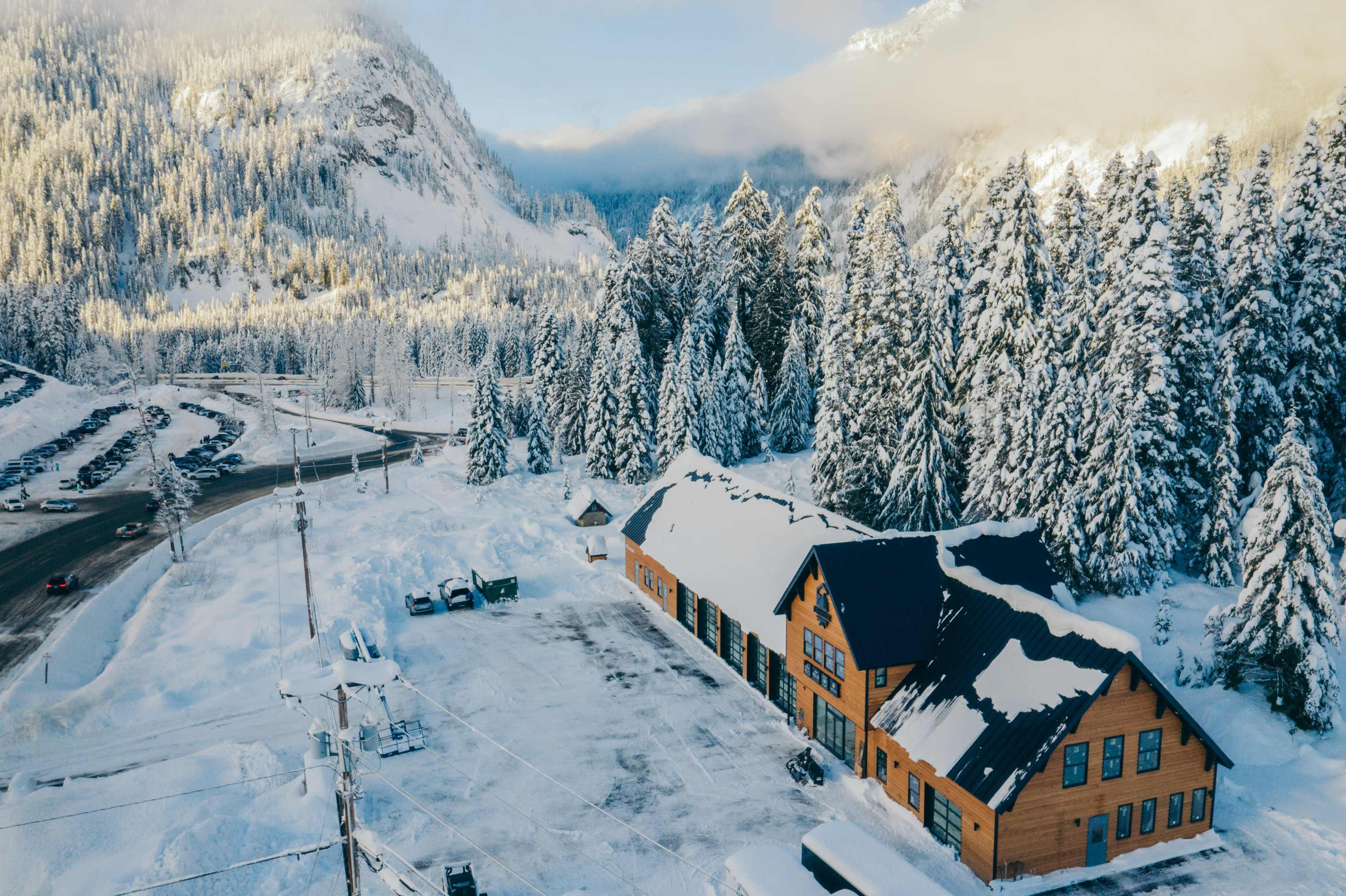 A snow-covered lodge is situated next to a forest of tall, evergreen trees with mountains in the background.