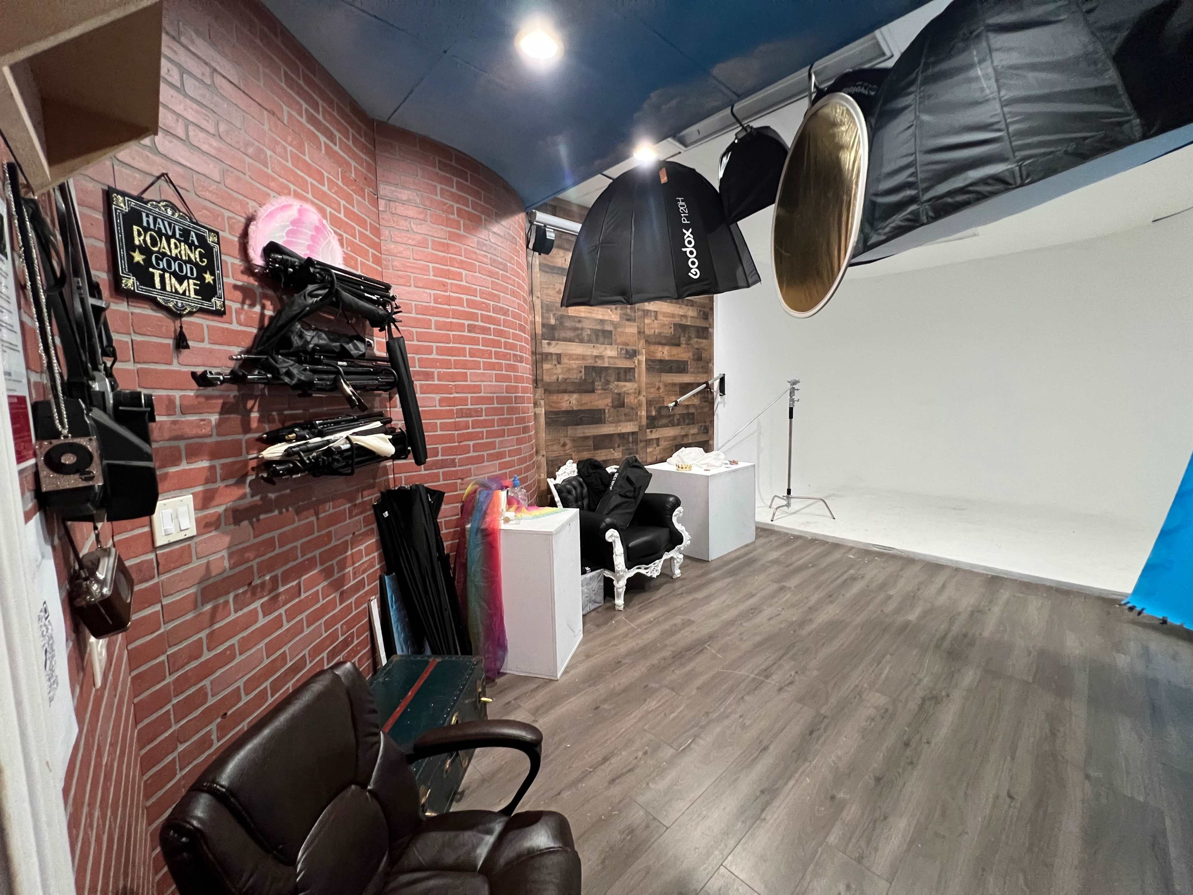 A photography studio with a brick wall, various lighting equipment hanging from the ceiling, and a white backdrop area.
