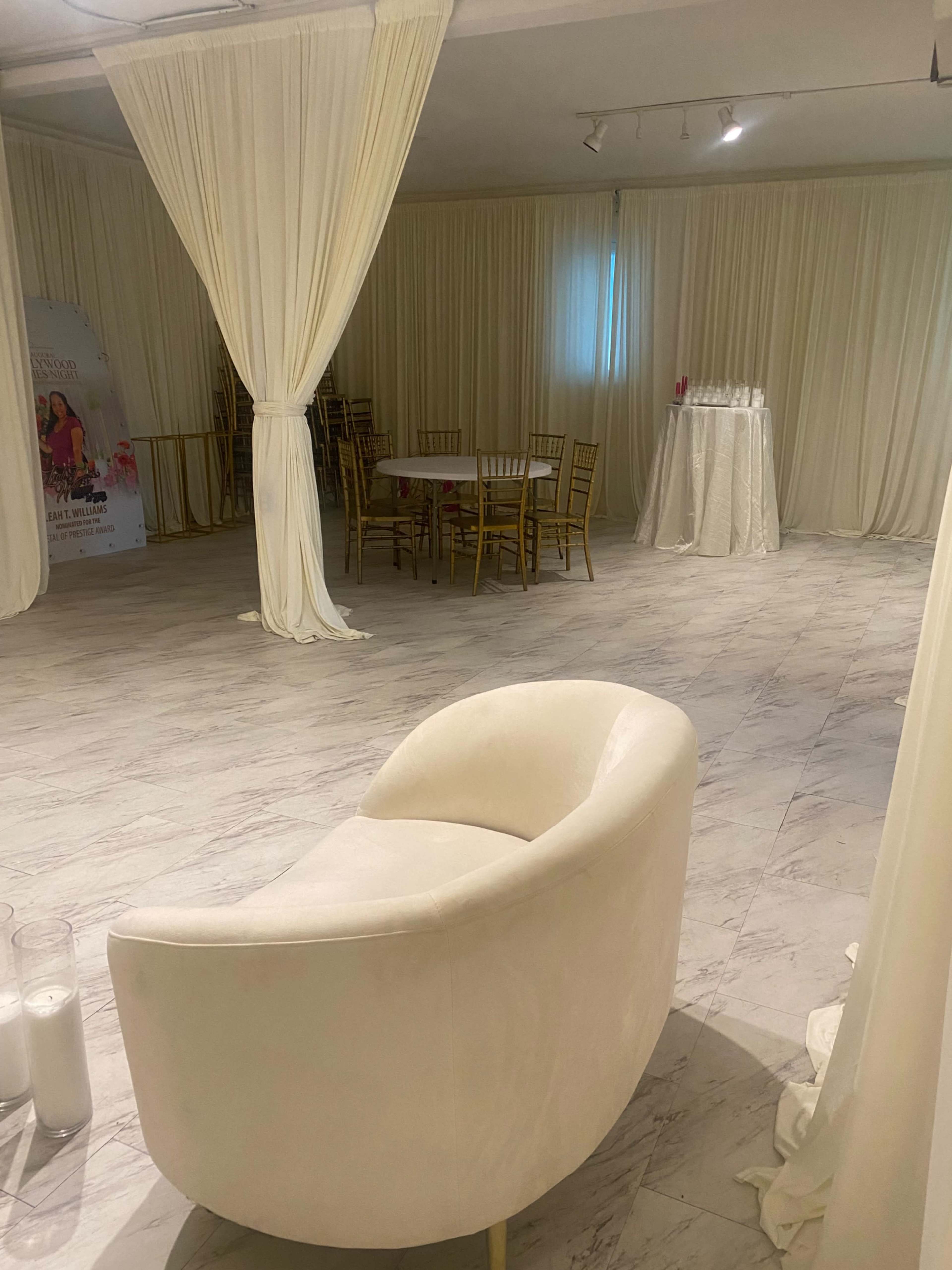 The image shows a spacious event venue decorated with white drapes and featuring a circular white chair, several tables with chairs, and a stand for drinks.