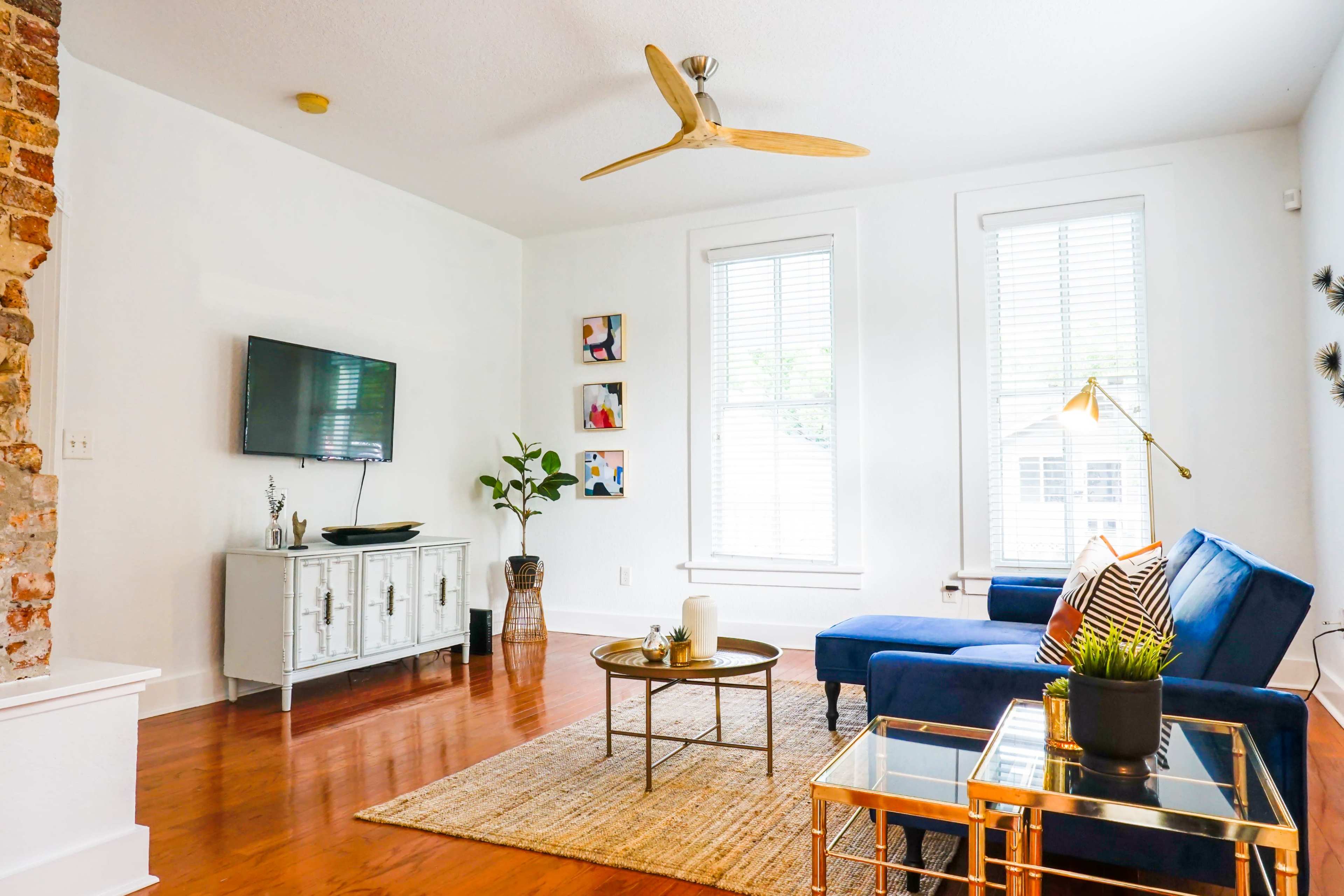 A modern living room with a blue sofa, a wooden coffee table, and a wall-mounted television, featuring large windows that allow natural light to enter.
