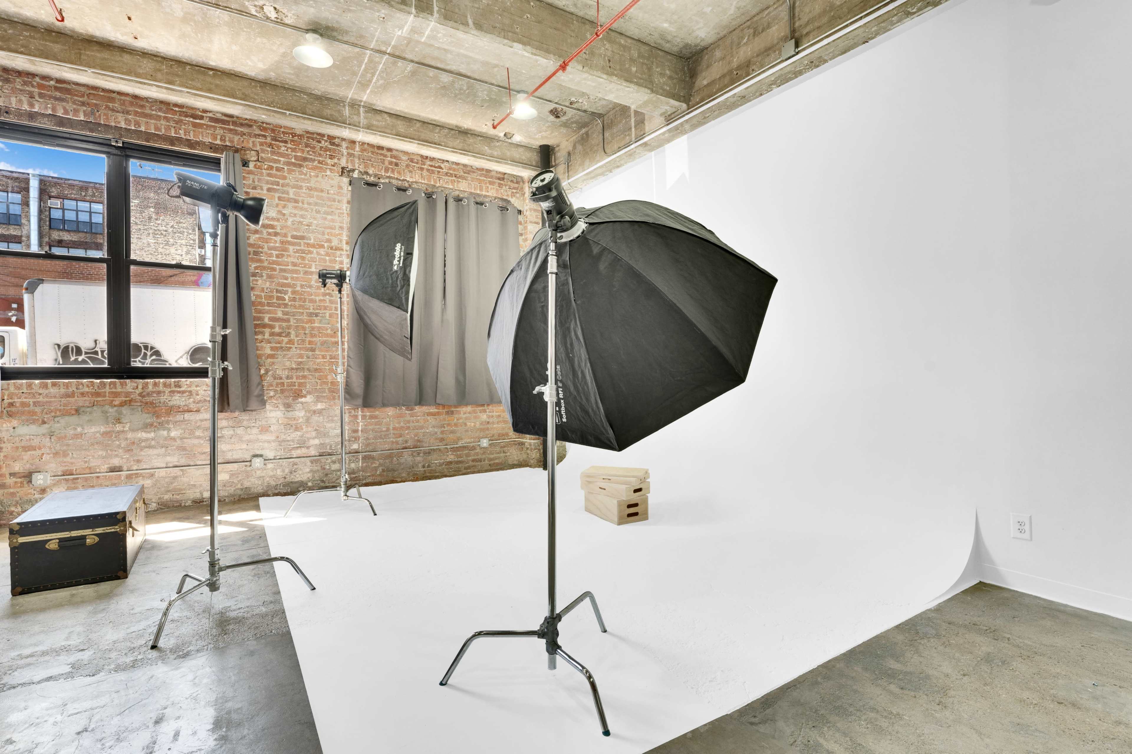 A photography studio featuring a white backdrop, two lighting stands with softboxes, and exposed brick walls.