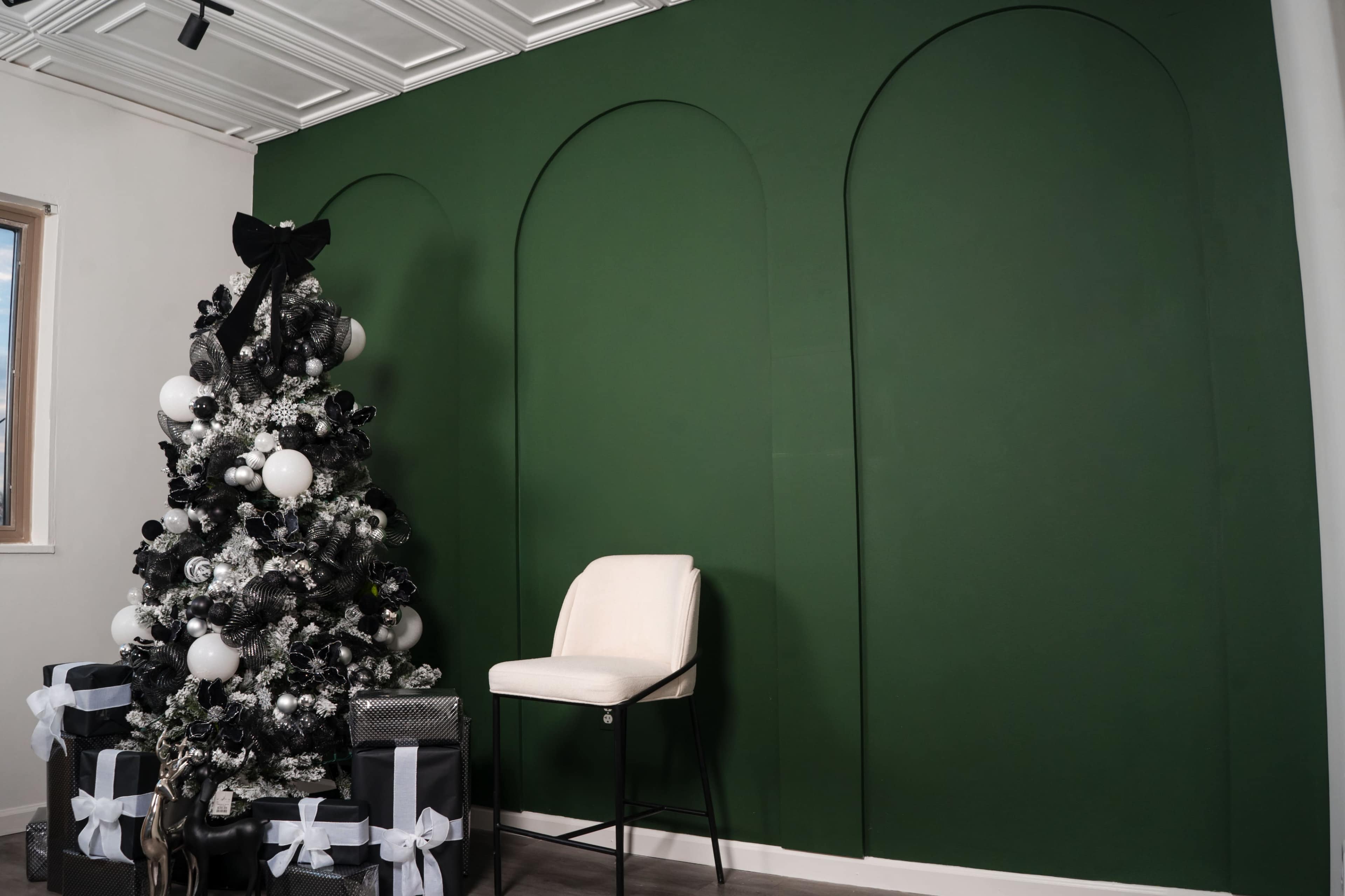 A decorated Christmas tree with black and white ornaments stands next to a modern white chair in front of a green curved wall.