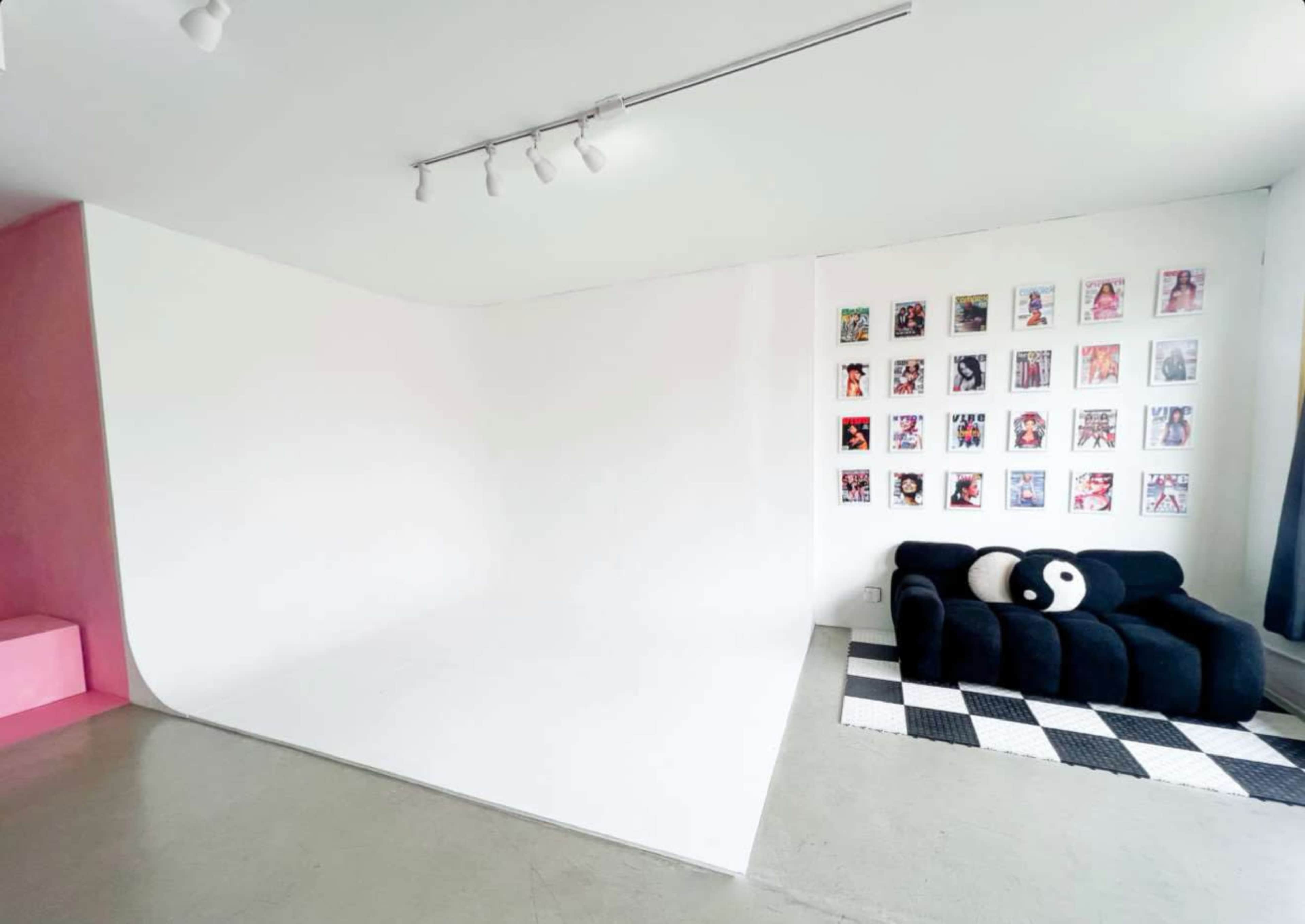 The image shows a minimalist studio space with a curved white backdrop, a black sofa adorned with a yin-yang pillow, and a wall displaying a gallery of colorful framed photographs.