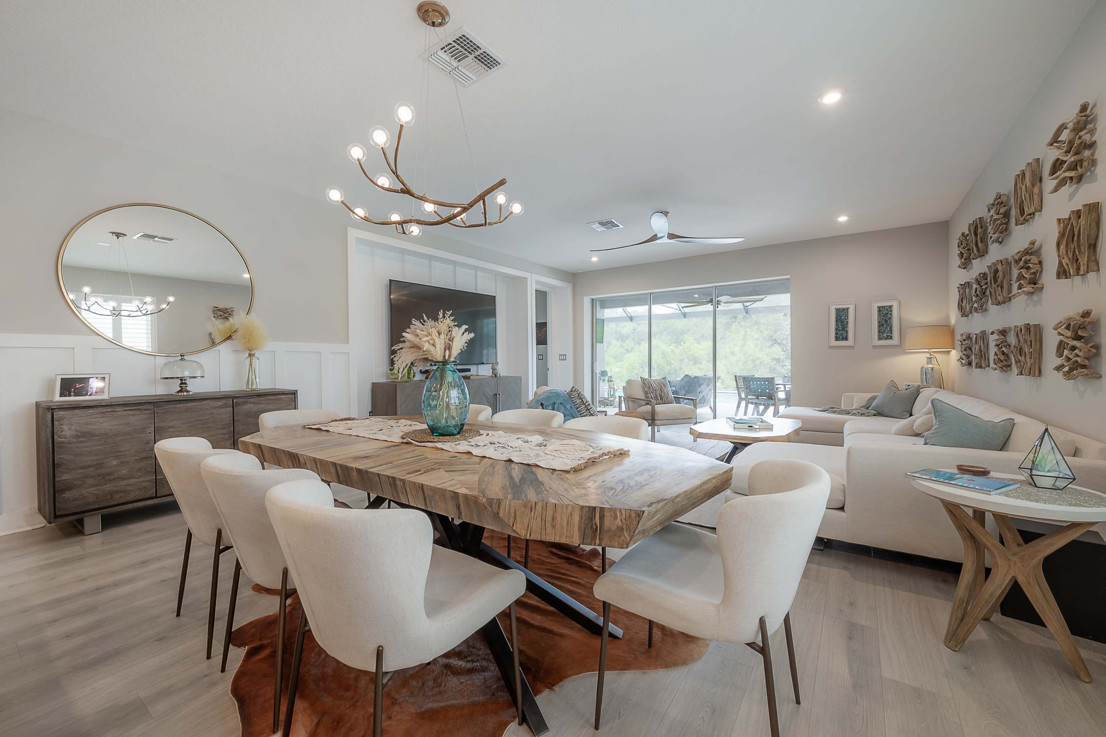 The image shows a modern dining area featuring a wooden table surrounded by upholstered chairs, with a living space in the background adorned with decorative wall art and contemporary lighting.
