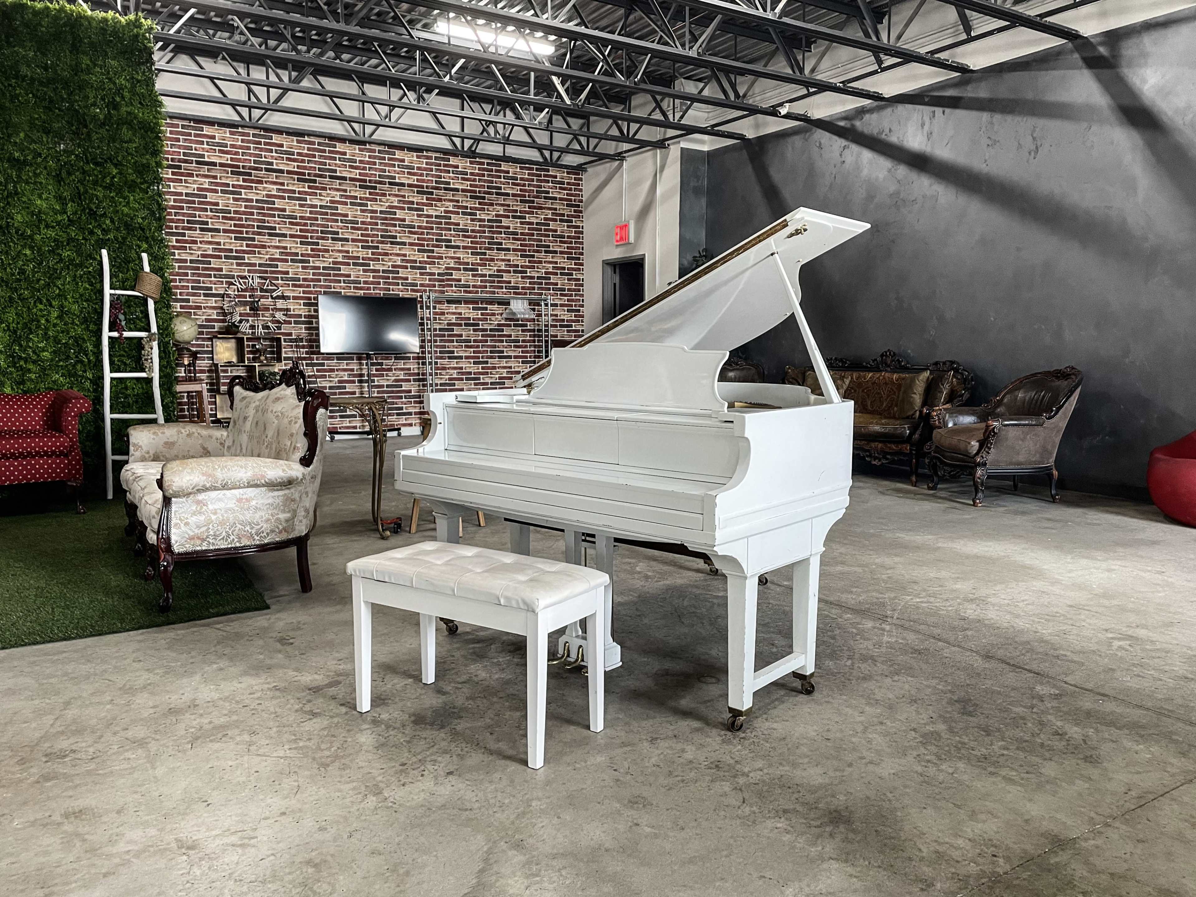 A white grand piano and matching bench are positioned centrally in a spacious industrial-style room with a brick wall and various vintage furniture pieces.