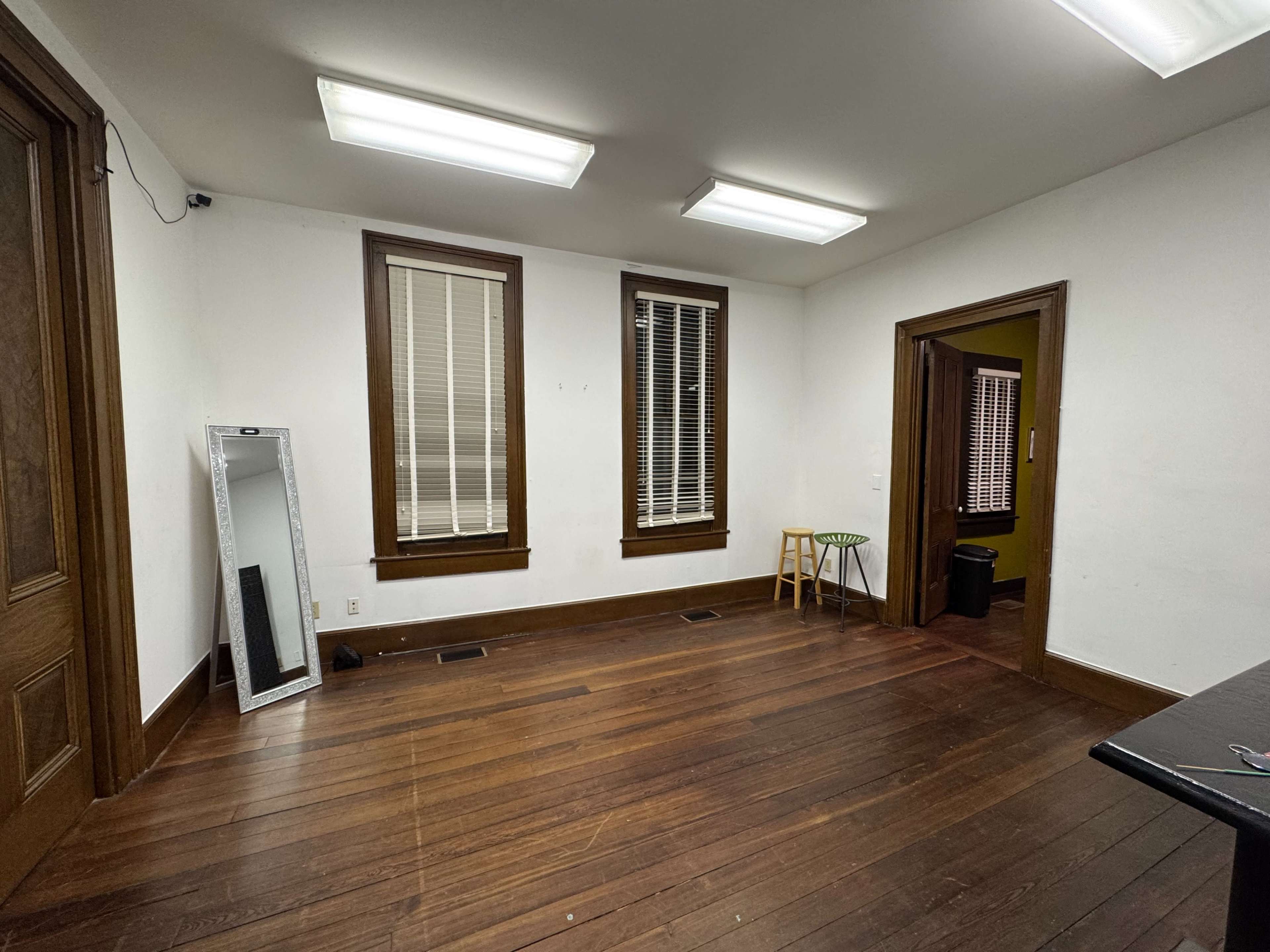 The image shows a sparsely furnished room with wooden flooring, two windows with blinds, and a mirror leaning against a wall.