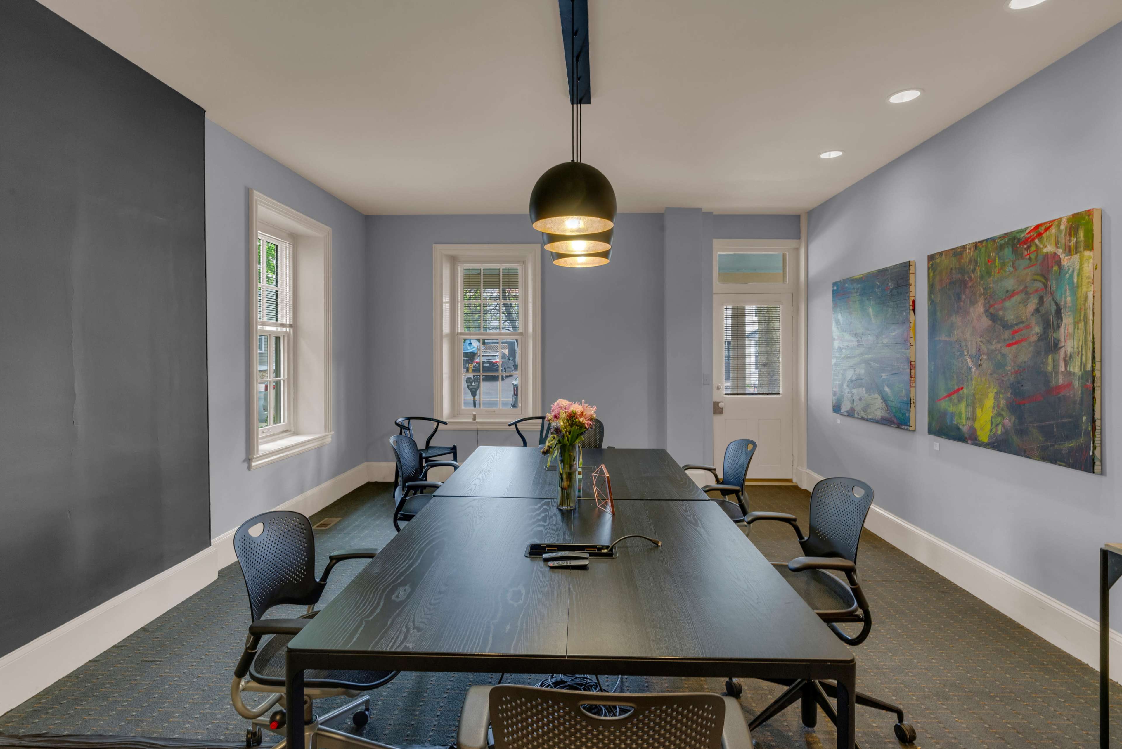 A conference room features a large black table surrounded by office chairs, with artwork on the walls and natural light coming in from the windows.