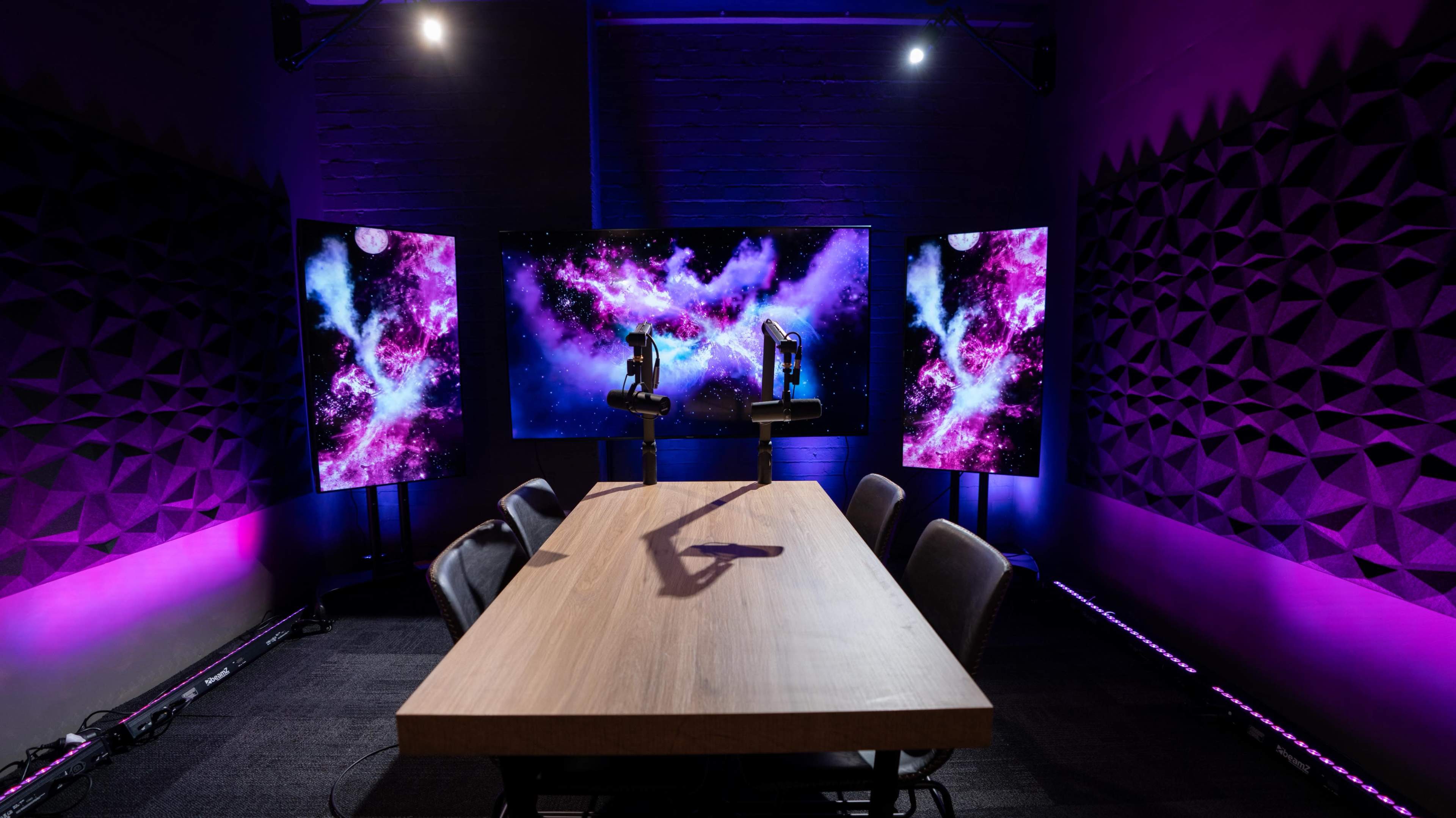 A conference room features a wooden table with black chairs, flanked by two large screens displaying a colorful cosmic background.