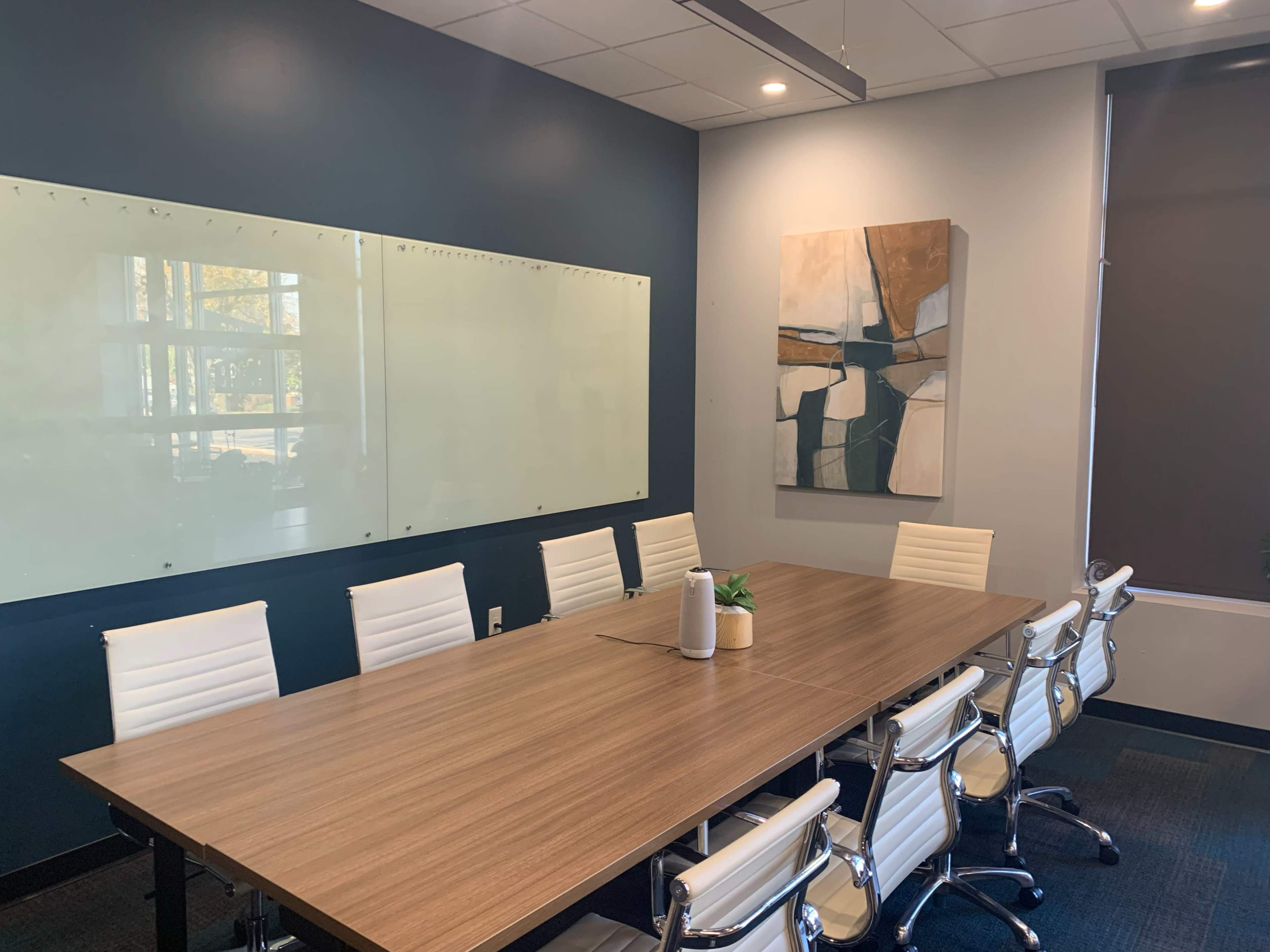 The image shows a modern conference room with a long wooden table, several white chairs, and a large whiteboard along one wall, complemented by an abstract painting on another wall.