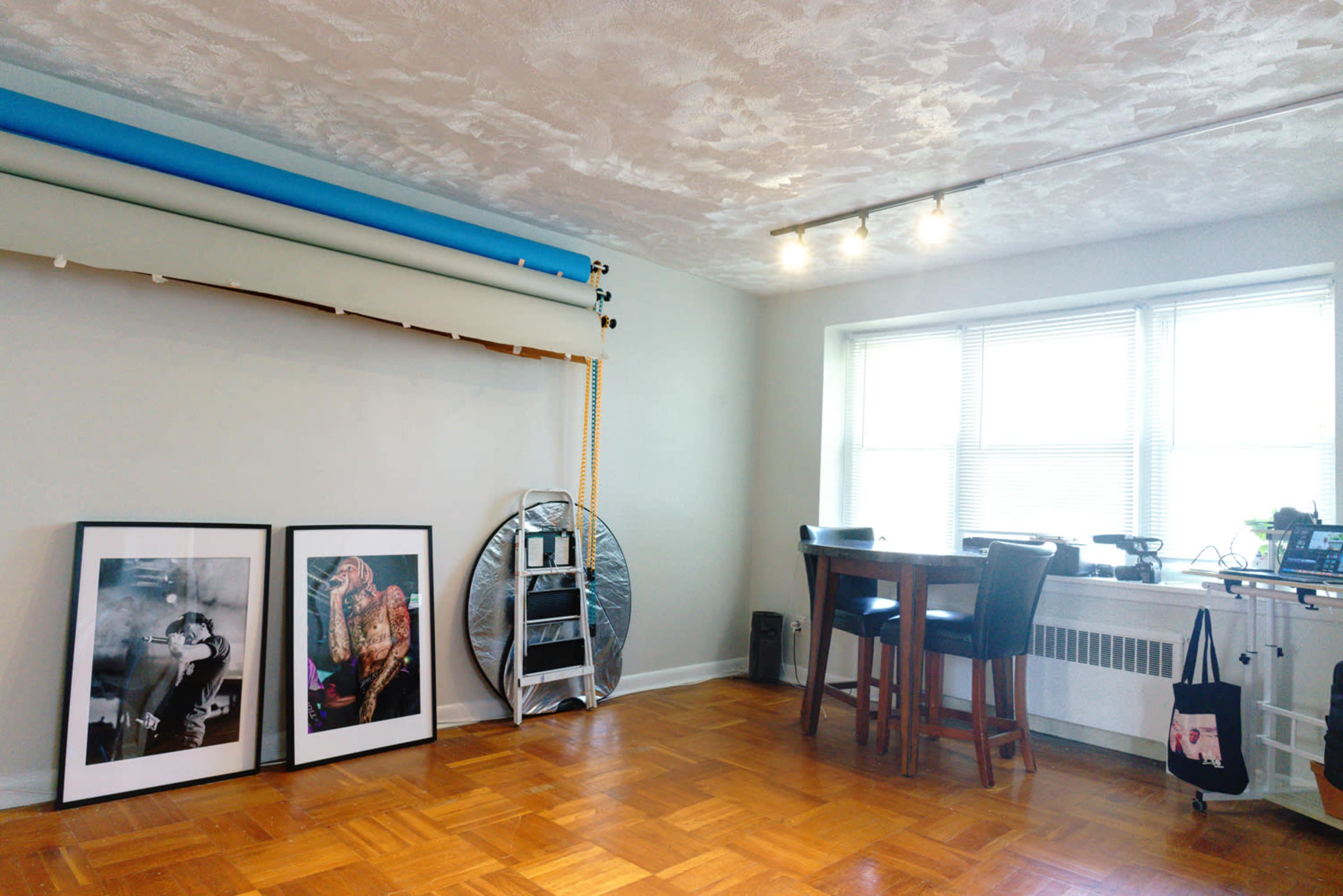 A minimalistic room featuring two framed photographs on the floor, a backdrop setup, a round table with chairs, and a window with multiple light sources.