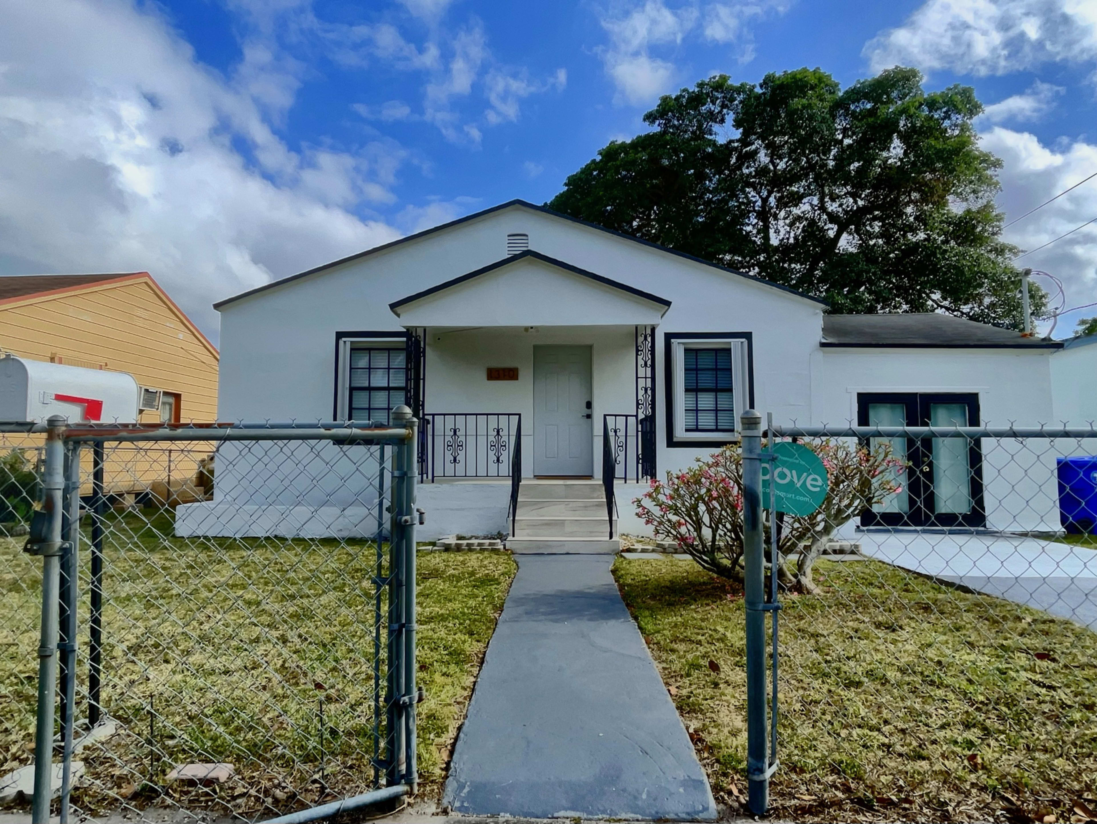 A white house with a front porch and a fenced yard is seen under a partly cloudy sky.