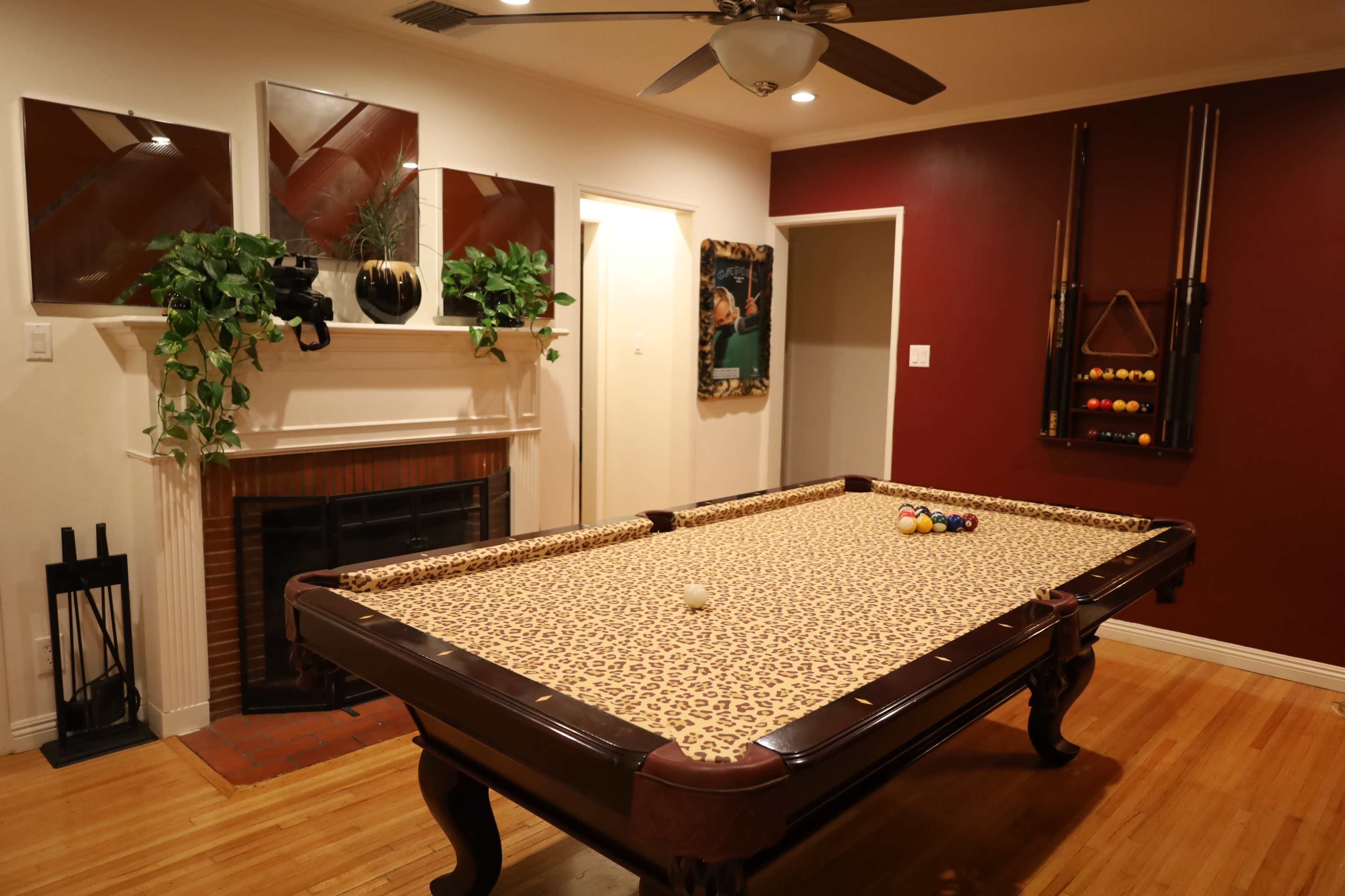 The image shows a game room featuring a leopard-print pool table, a fireplace, and decorative wall mirrors, complemented by a red accent wall and a rack of pool cues.