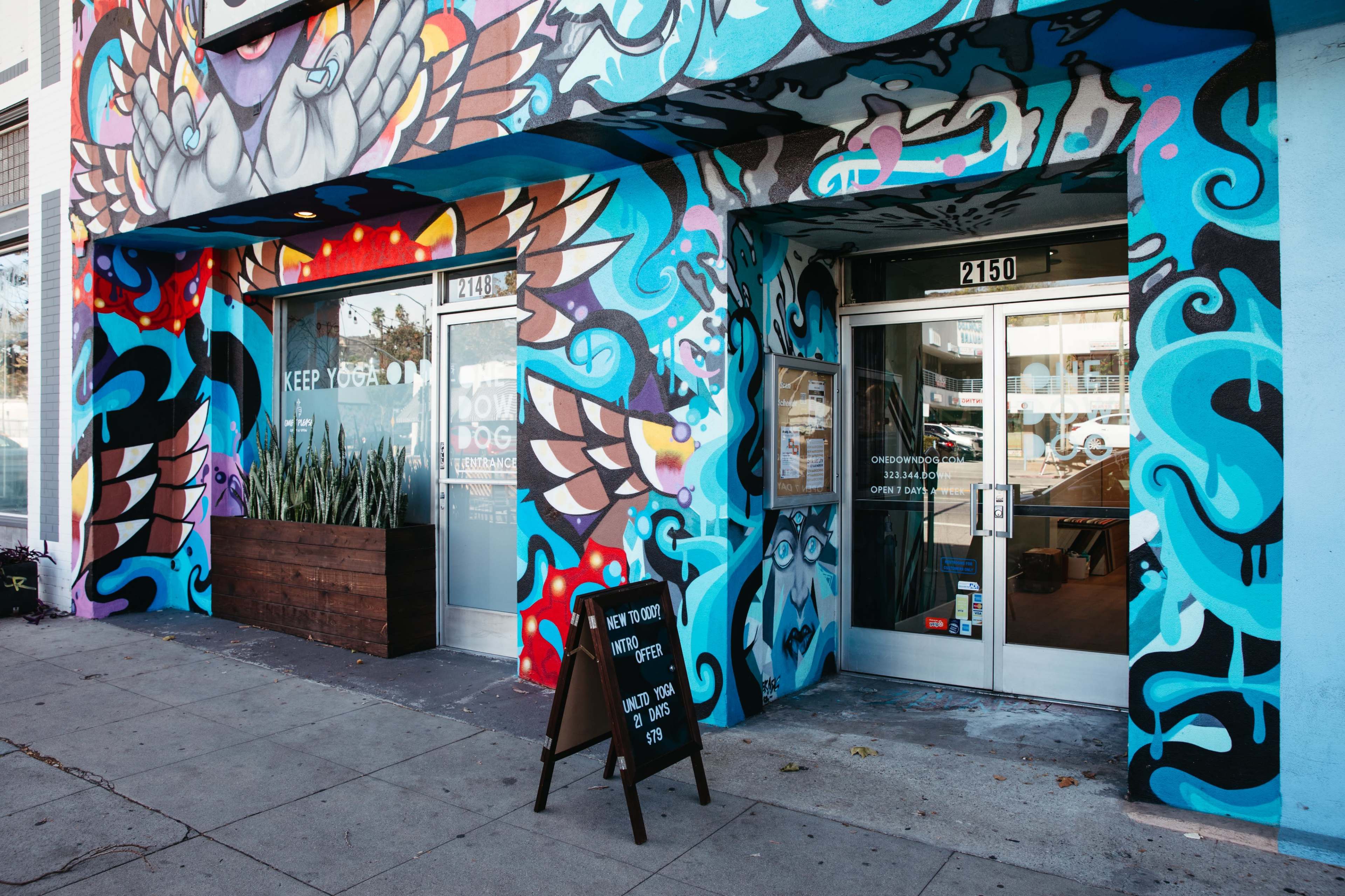 A colorful mural on a building facade with two glass entrance doors and a chalkboard sign outside.