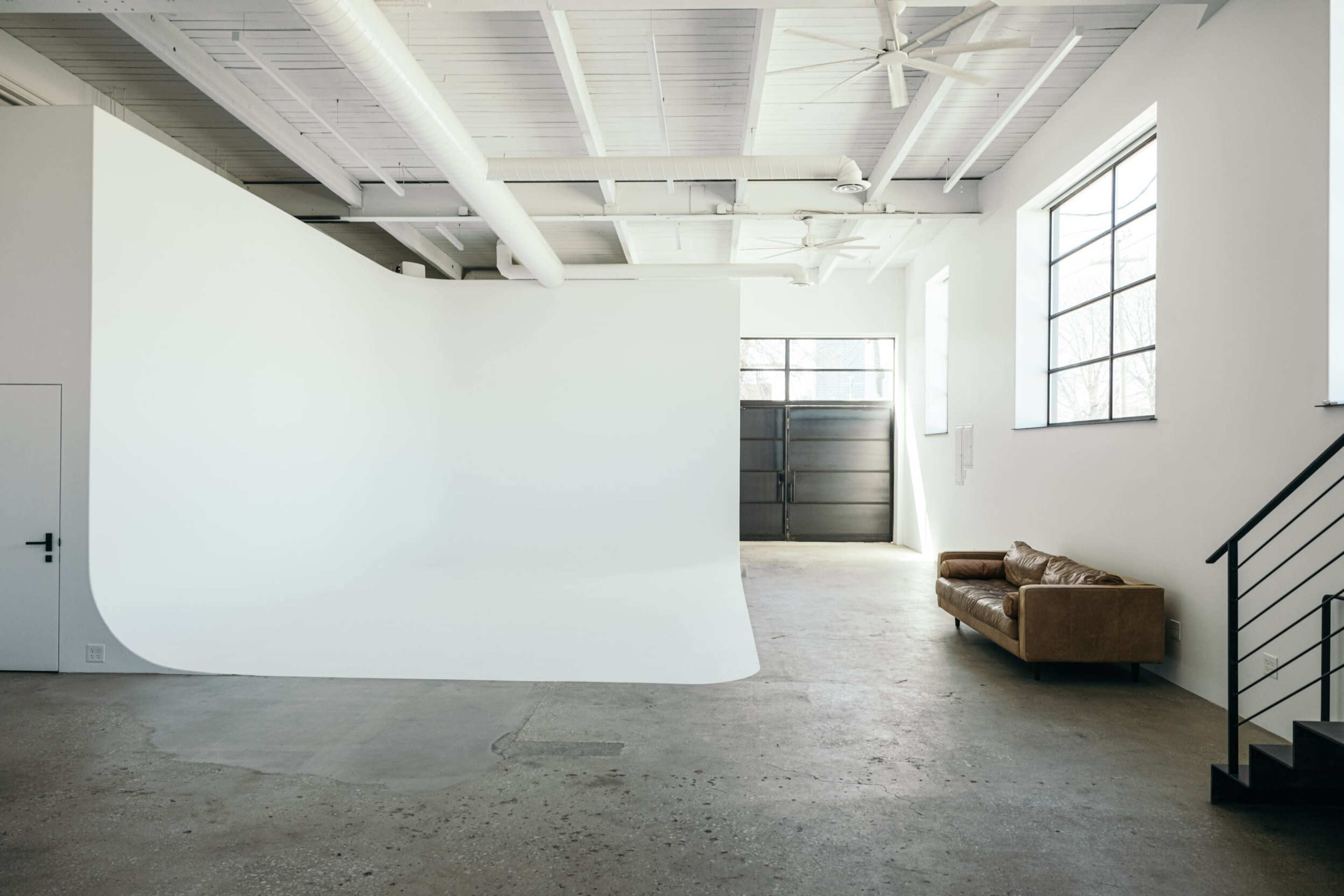 A large, empty studio space features a seamless white backdrop and a brown leather couch against a concrete floor.