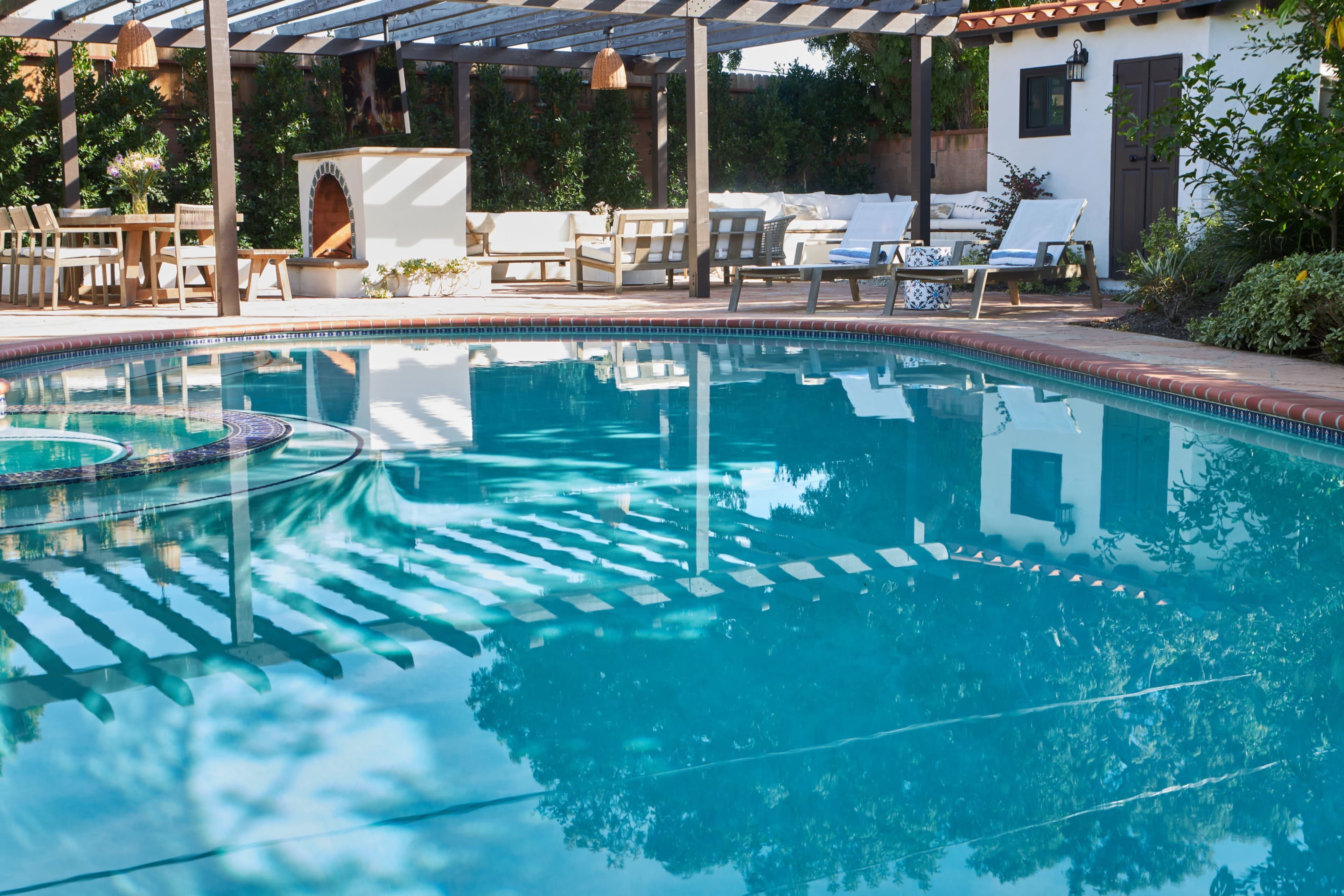 The image shows a tranquil pool area with a reflection of a pergola and outdoor seating surrounding a clear blue pool.