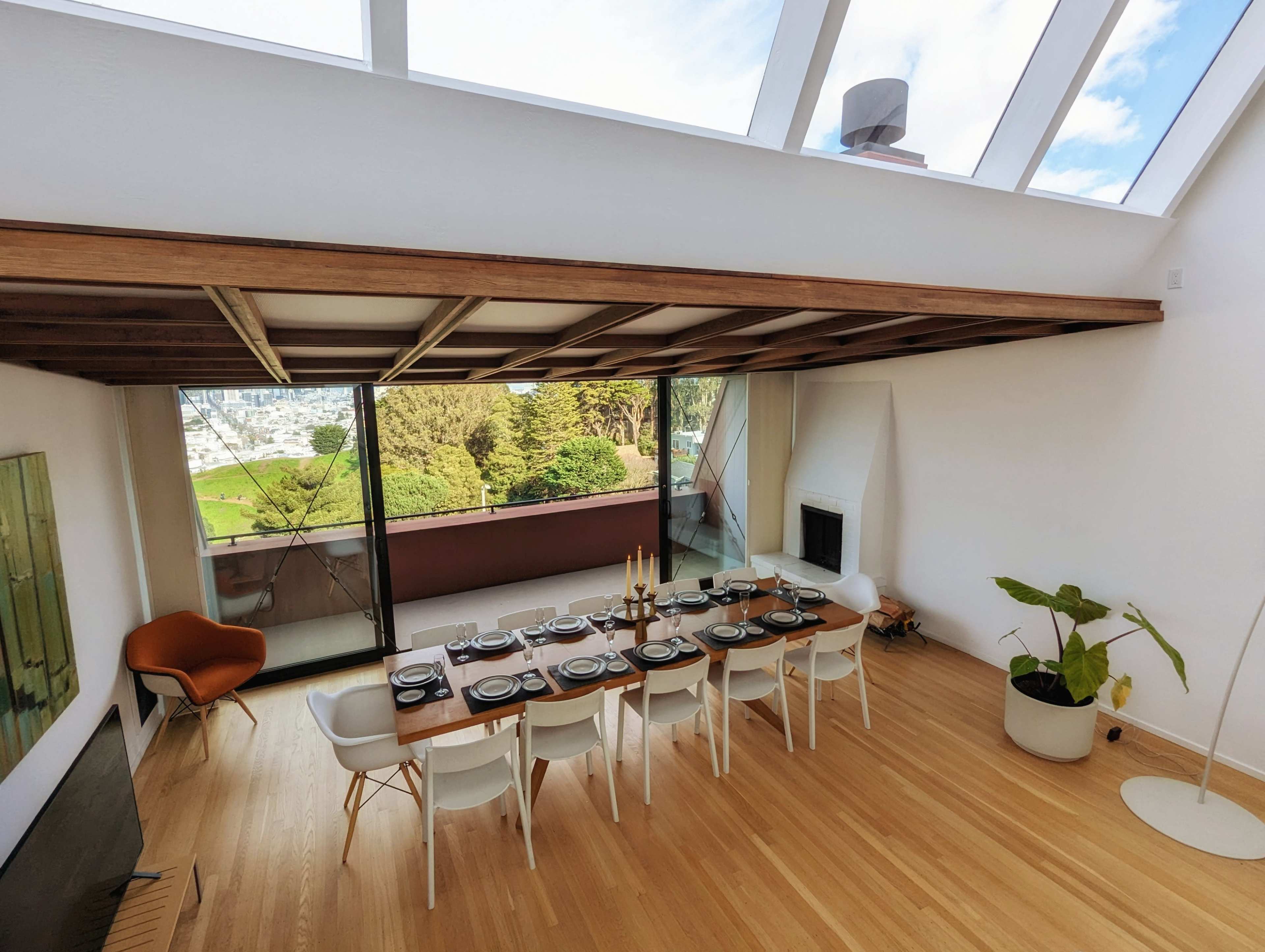 The image shows a dining room with a long wooden table set for a meal, surrounded by white chairs and large windows that provide a view of lush greenery outside.