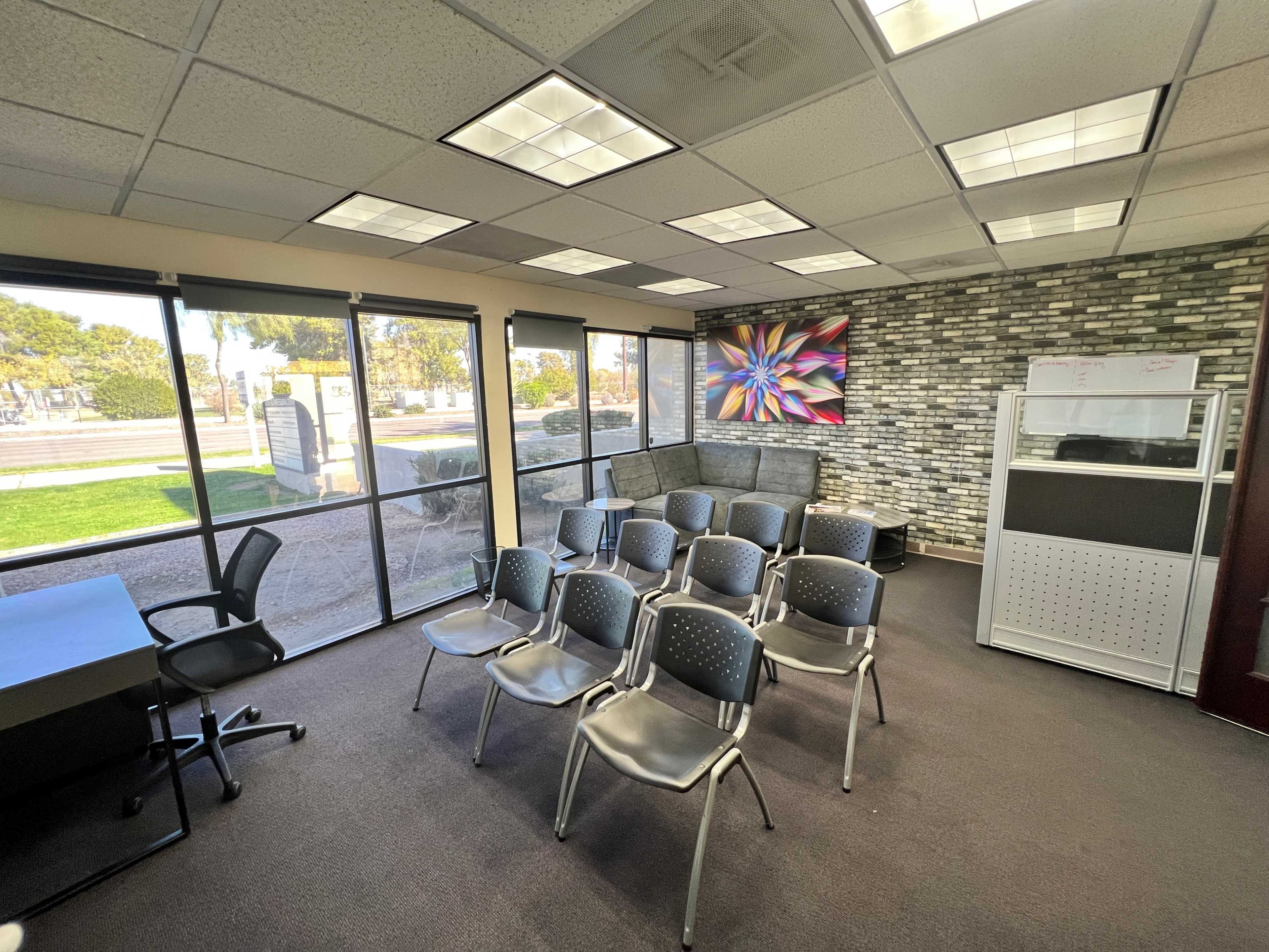 A conference room with several gray chairs arranged in front of a wall featuring a colorful artwork, alongside windows that provide outdoor views.