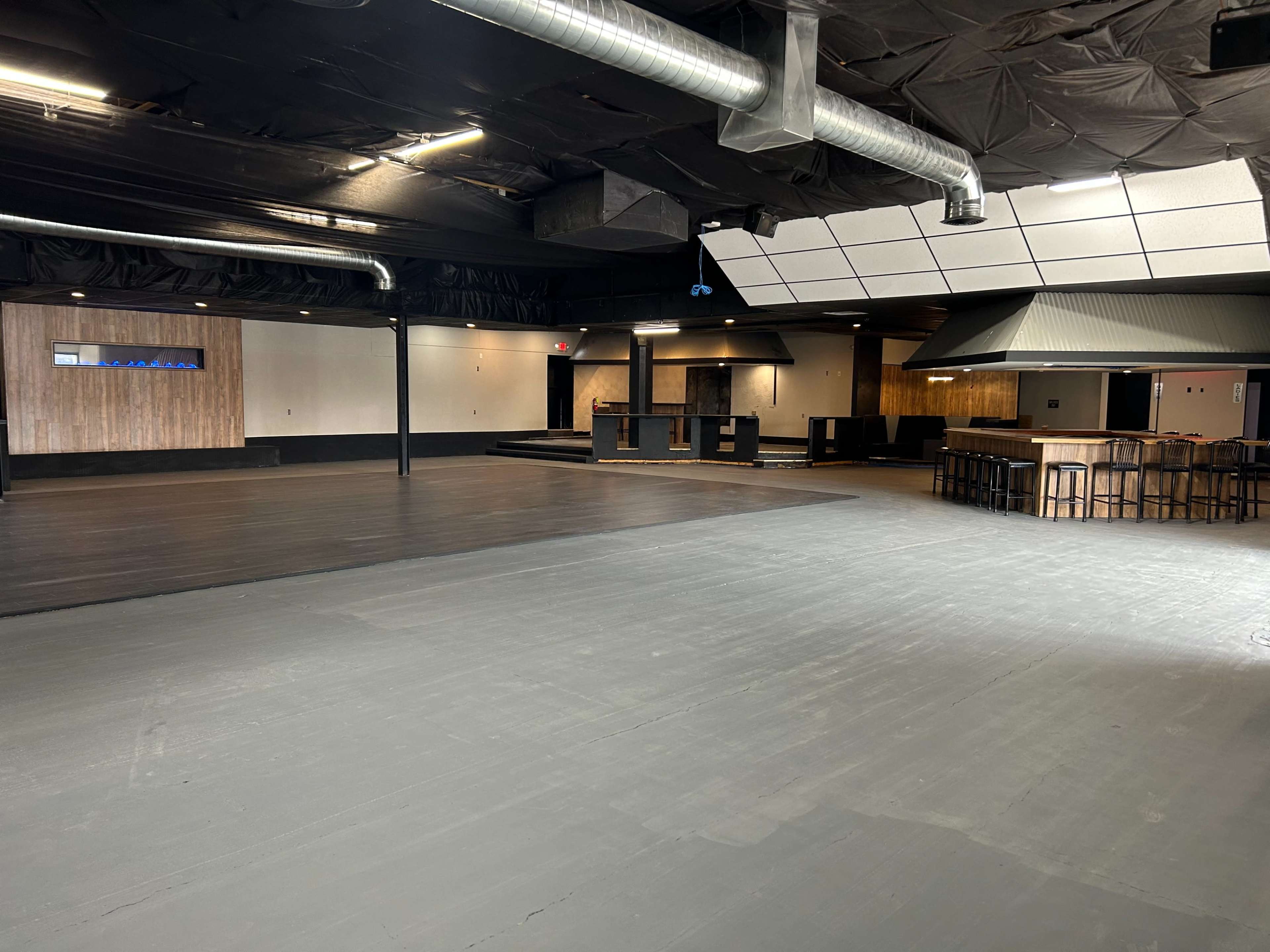 An empty indoor venue features a dark wooden stage and a bar area, with polished floors and exposed ductwork.