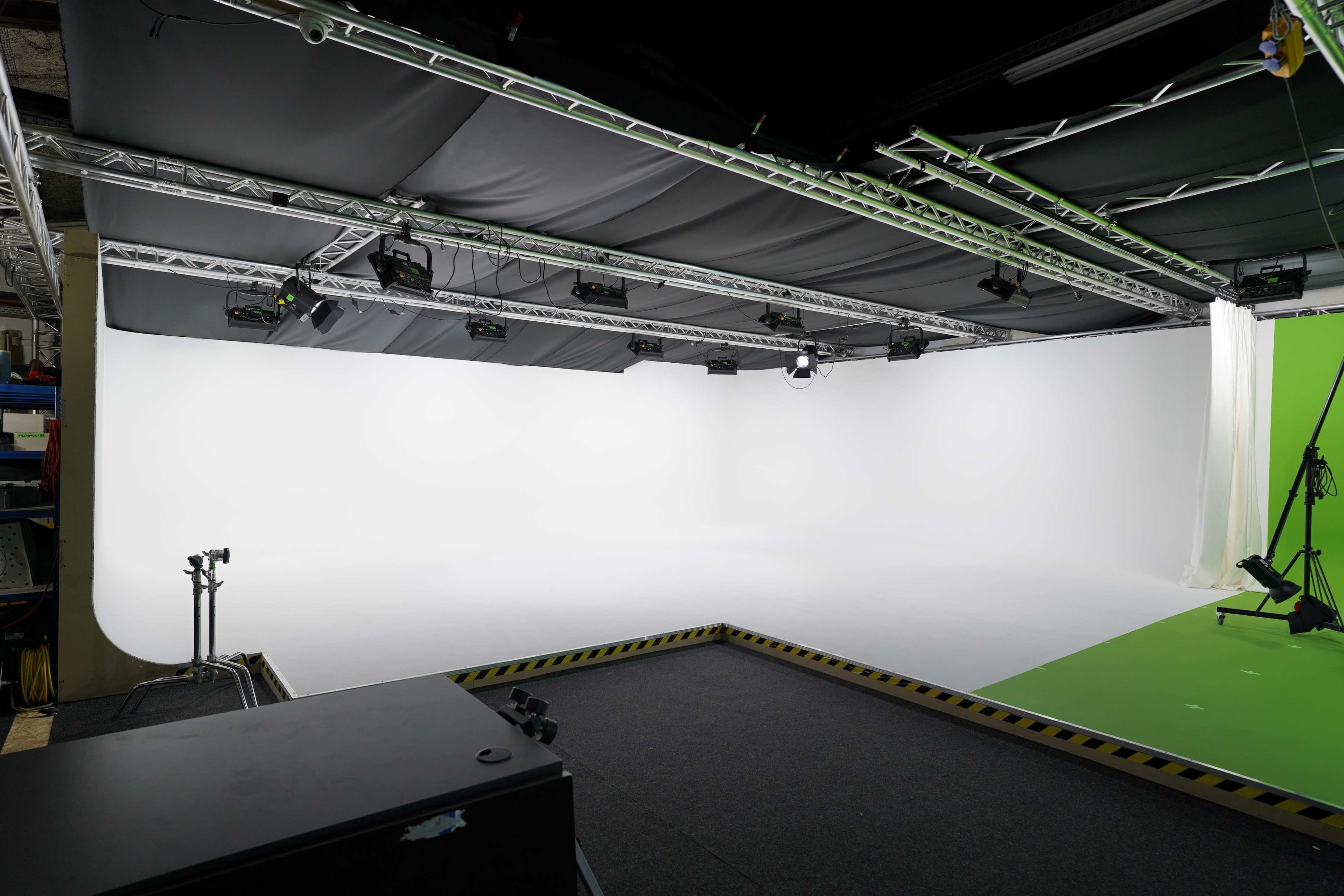 The image shows an empty studio space with a white background on one side and a green screen on the other, equipped with overhead lighting and cameras.