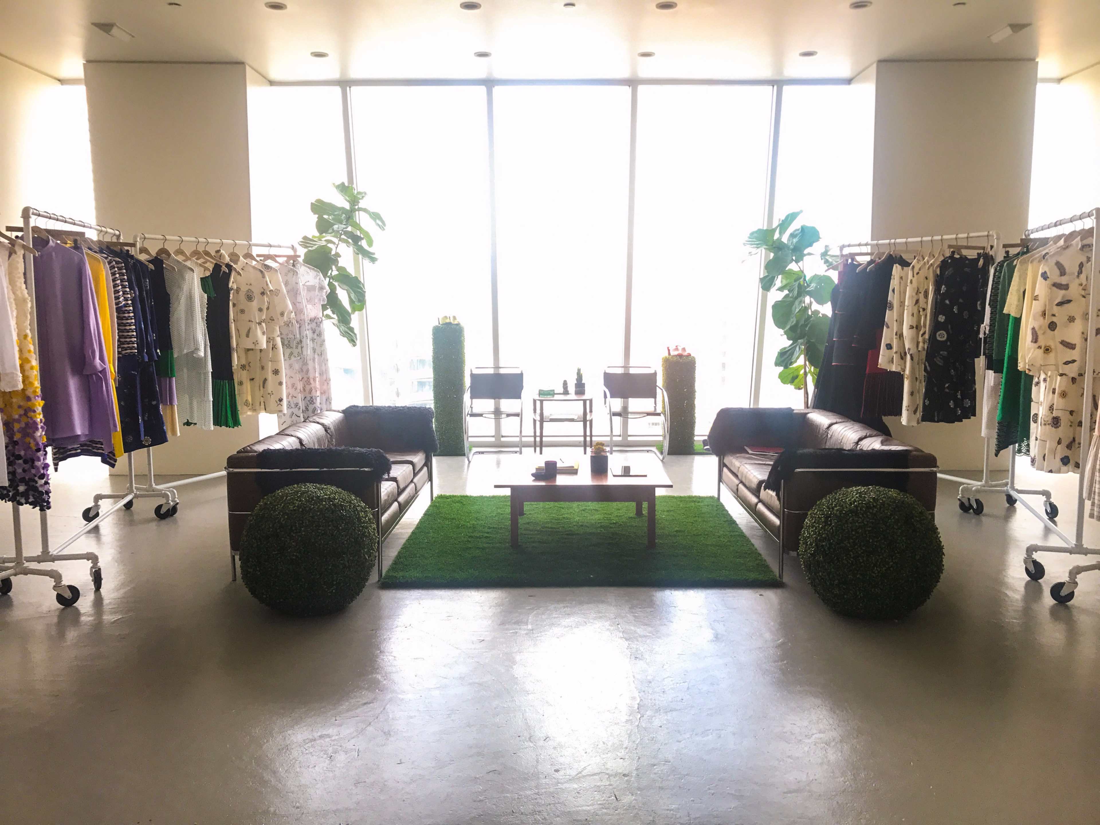 A spacious, well-lit room with two leather sofas facing a coffee table, surrounded by clothing racks and potted plants.