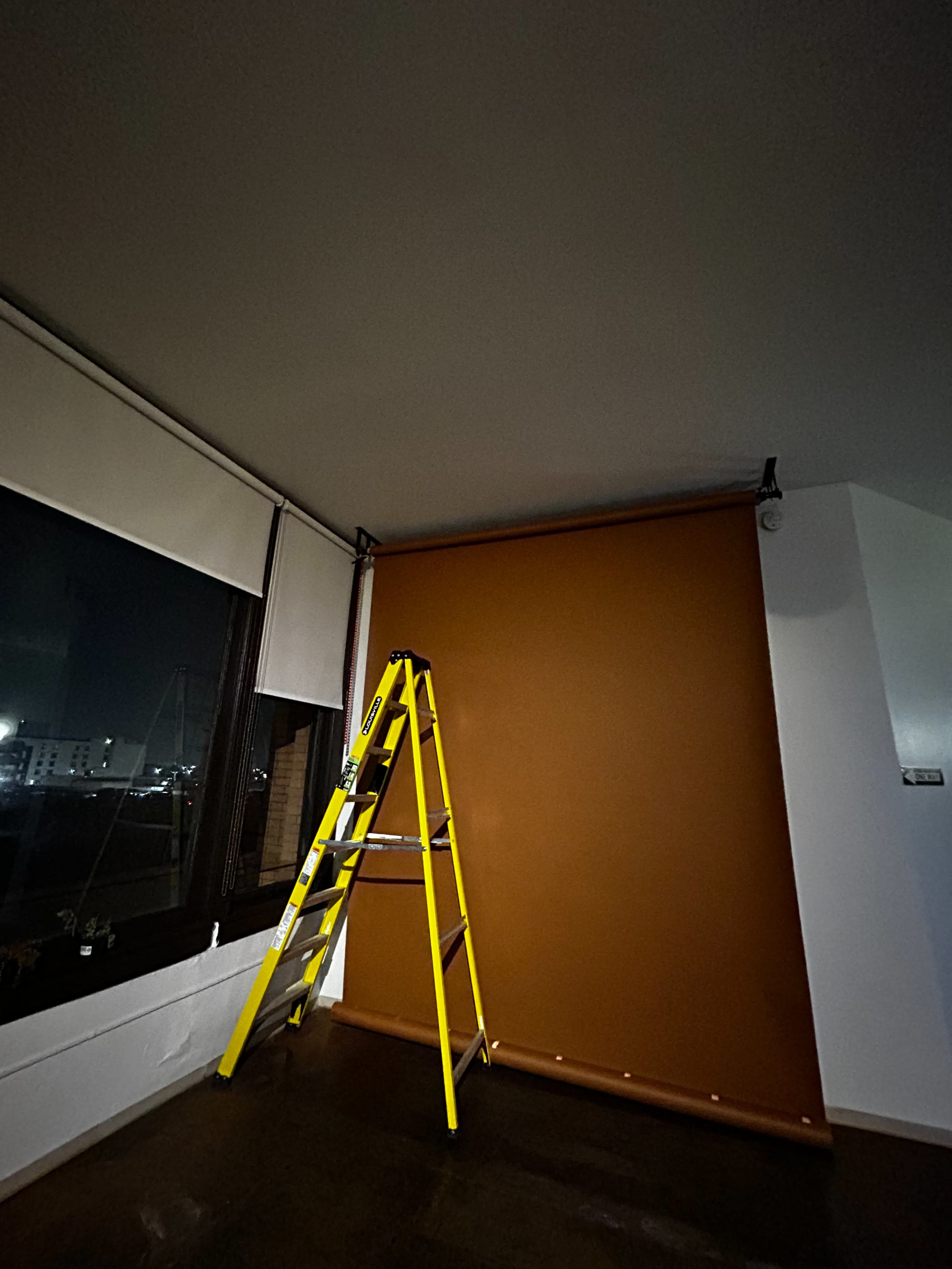 A yellow ladder stands next to a large brown roller shade that is mounted to the wall in a dimly lit room.