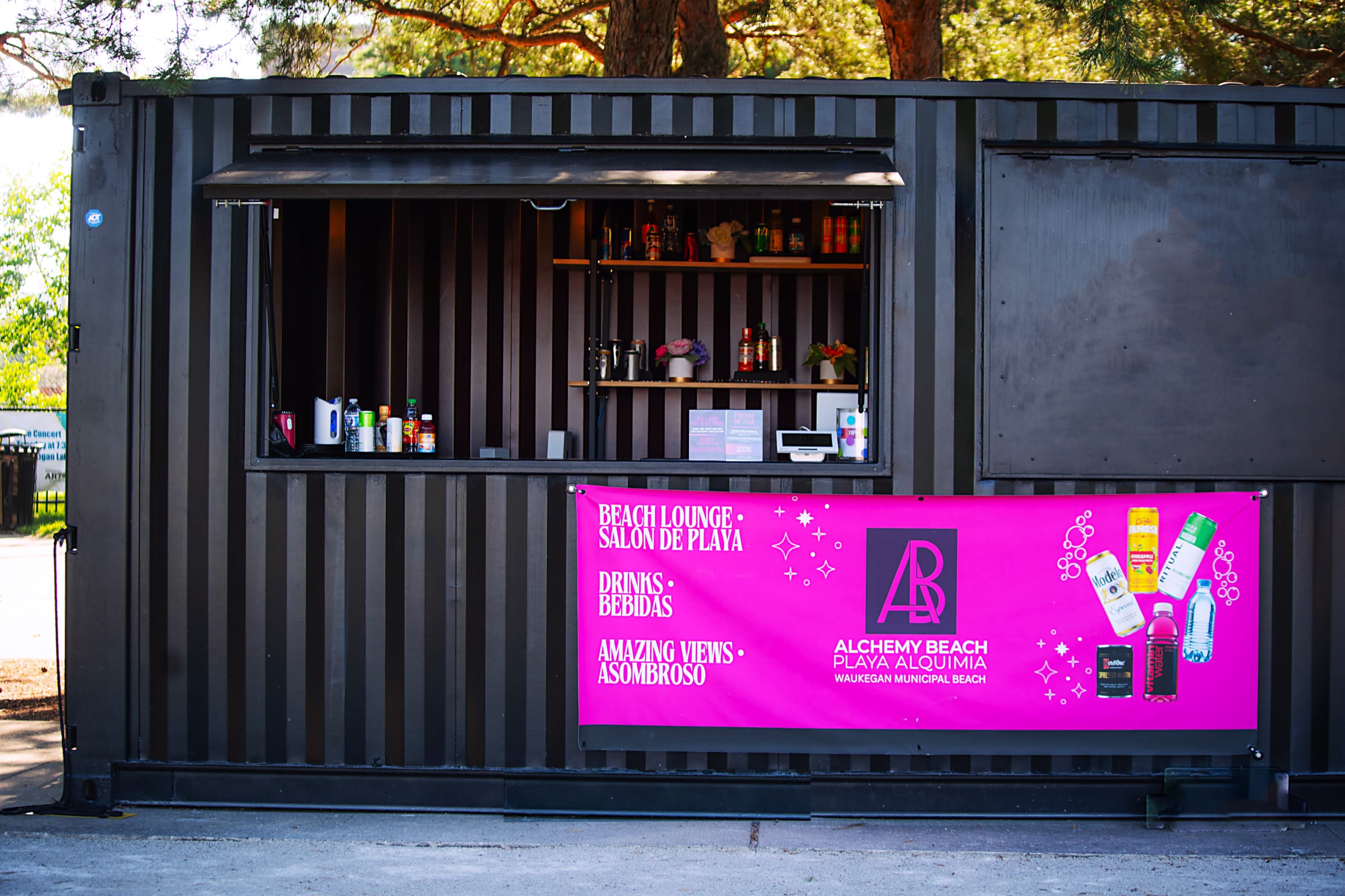 The image displays a beach bar constructed from a shipping container, featuring a menu of drinks and a vibrant sign showcasing the bar's name and offerings.