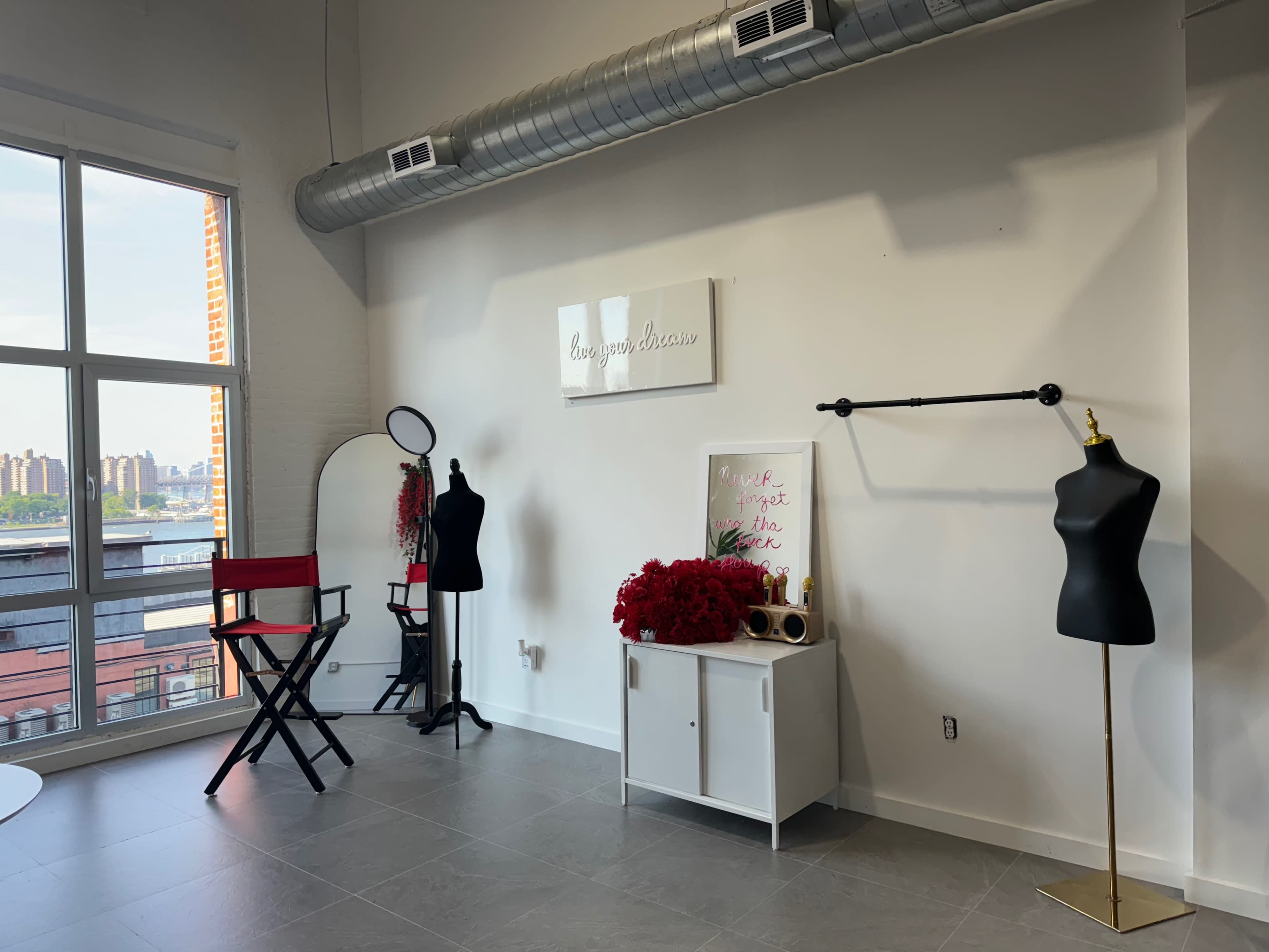 A spacious studio with large windows, featuring a red director's chair, two dress forms, a decorative sign on the wall, and a white storage unit adorned with red floral arrangements.