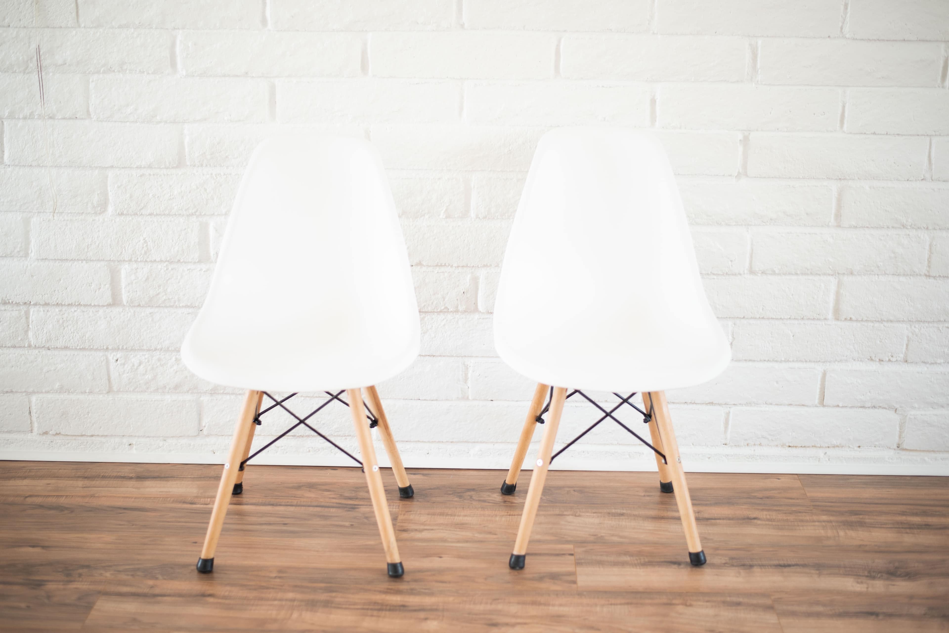 Two white plastic chairs with wooden legs are positioned back-to-back against a white brick wall.