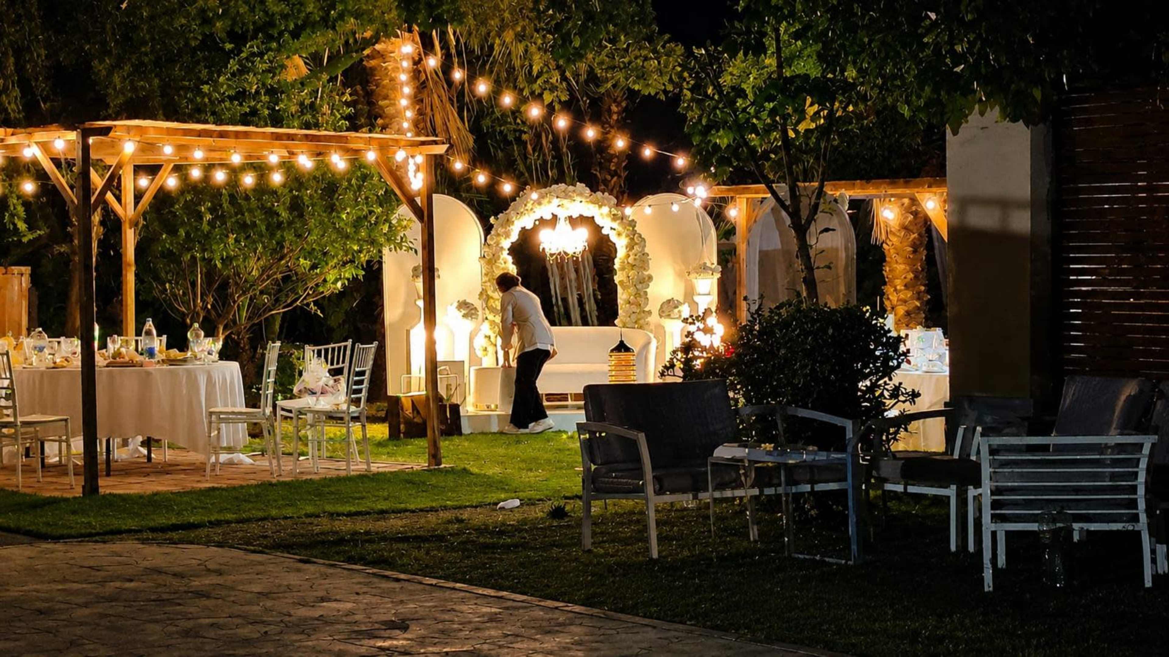 A garden party setup is illuminated by string lights, featuring decorated tables and a floral arch in the background.
