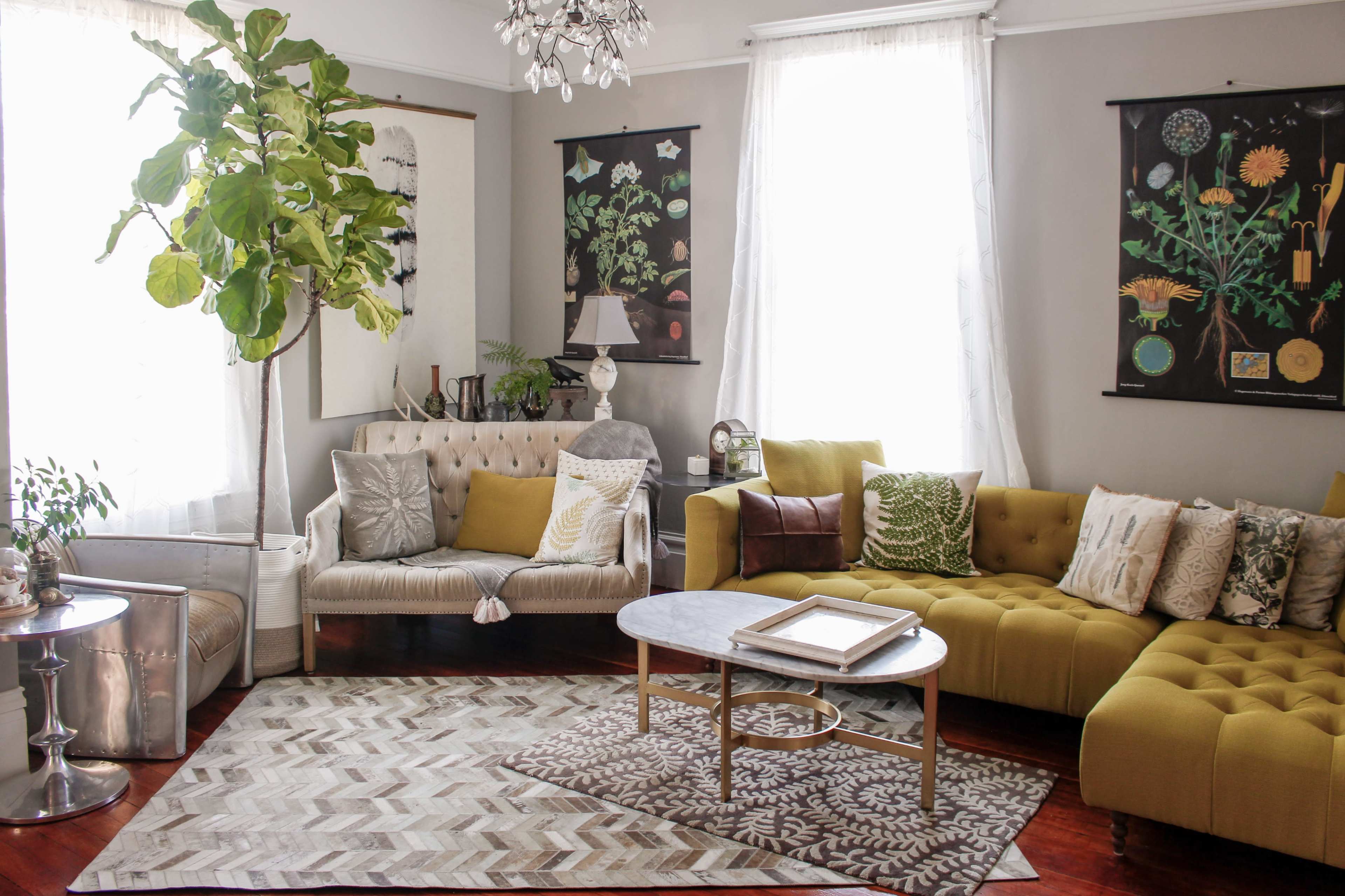 A bright living room features a mix of beige and green sofas, a round coffee table, and plants, with large windows allowing natural light to fill the space.