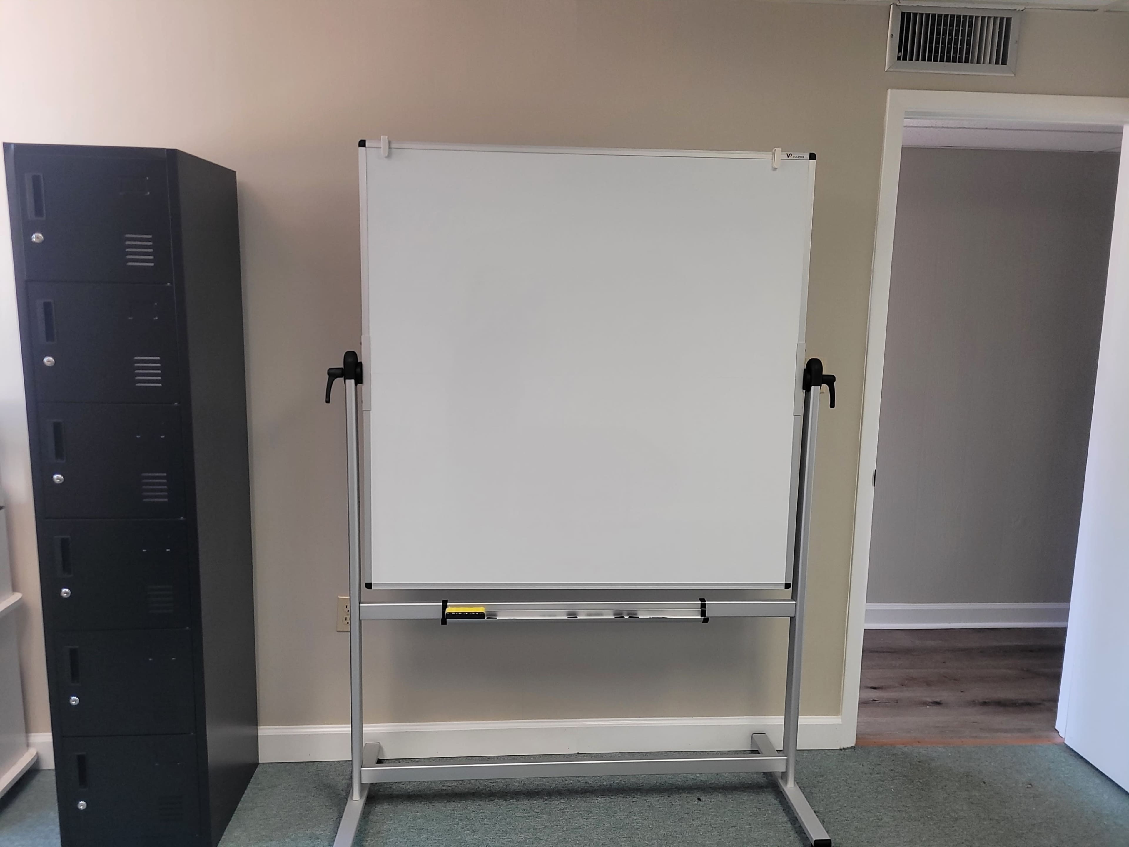 A whiteboard stands on a rolling easel in an office, next to a black storage cabinet and a doorway.