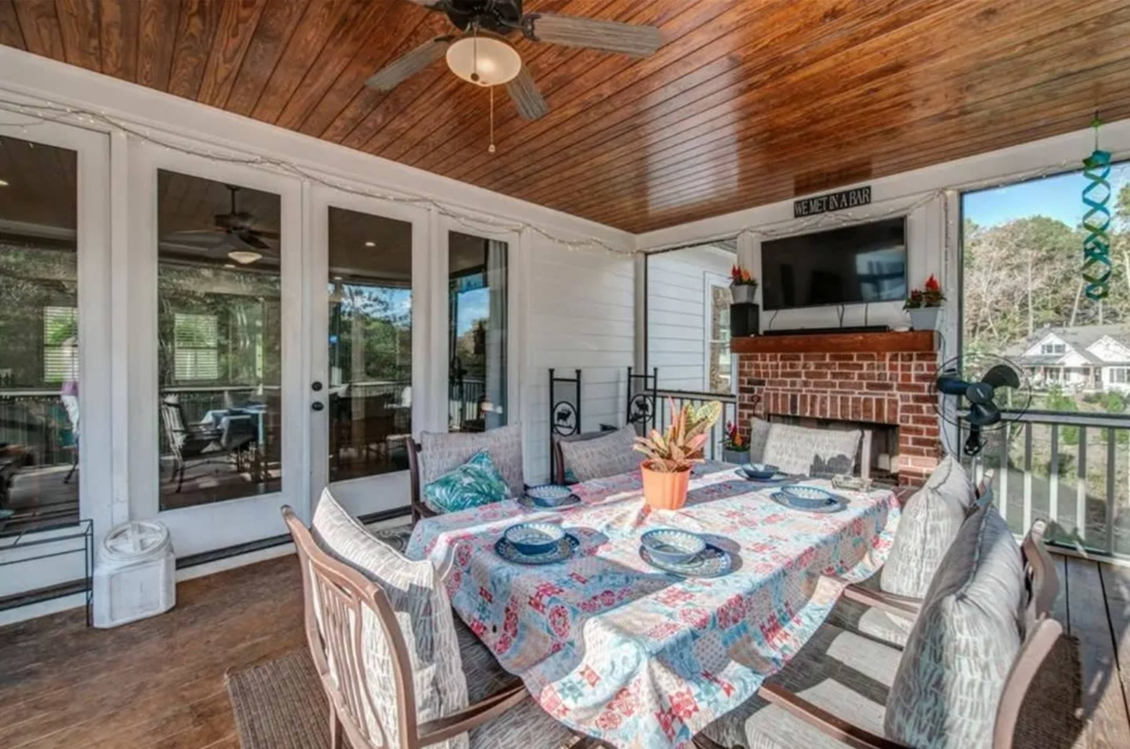 A screened patio features a dining table set with a colorful tablecloth, surrounded by chairs and a brick fireplace, with large windows offering a view of the outdoors.
