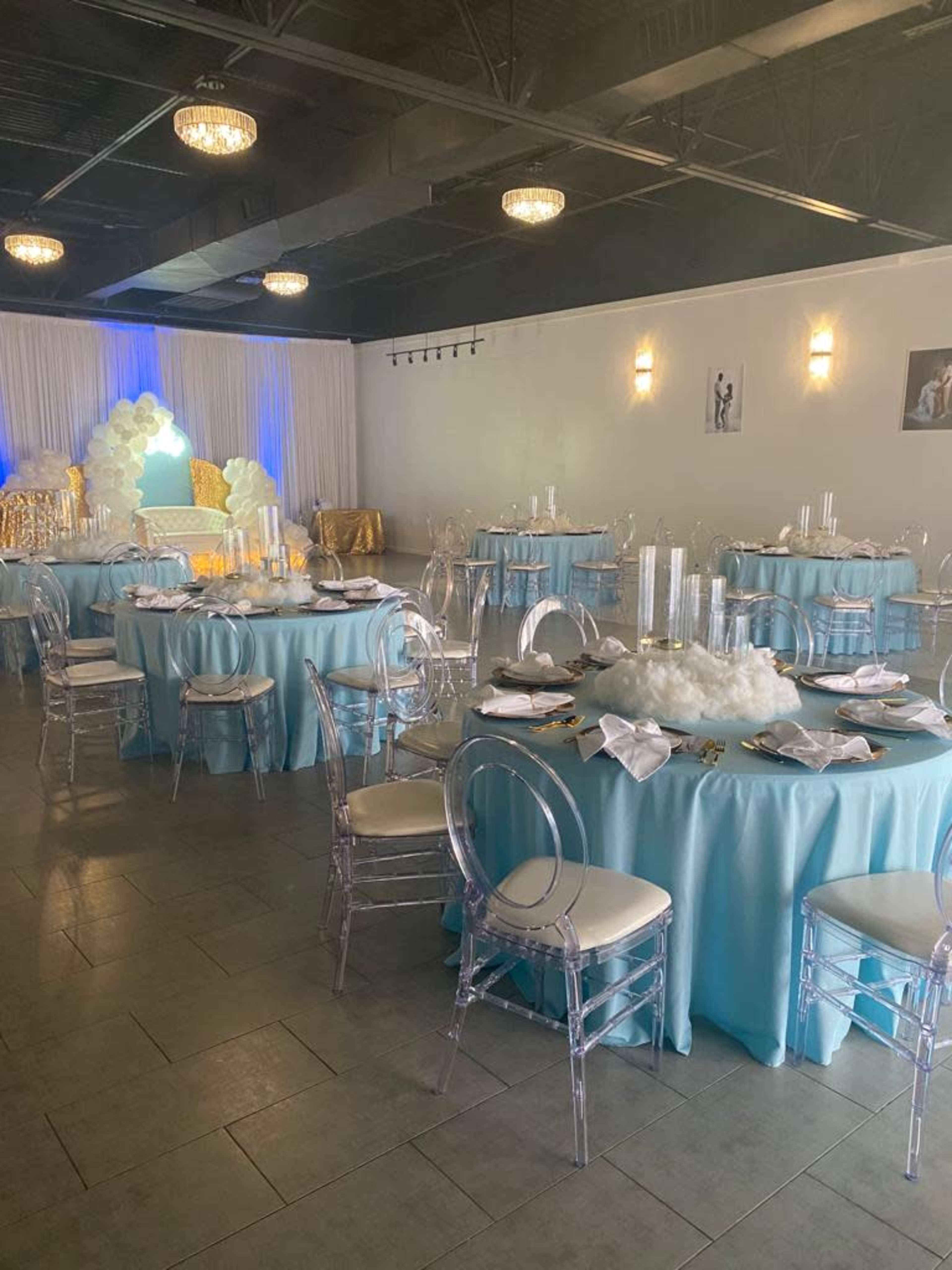 The image shows a decorated banquet hall featuring turquoise tablecloths, clear chairs, and a backdrop with white drapery and cloud-like decorations.