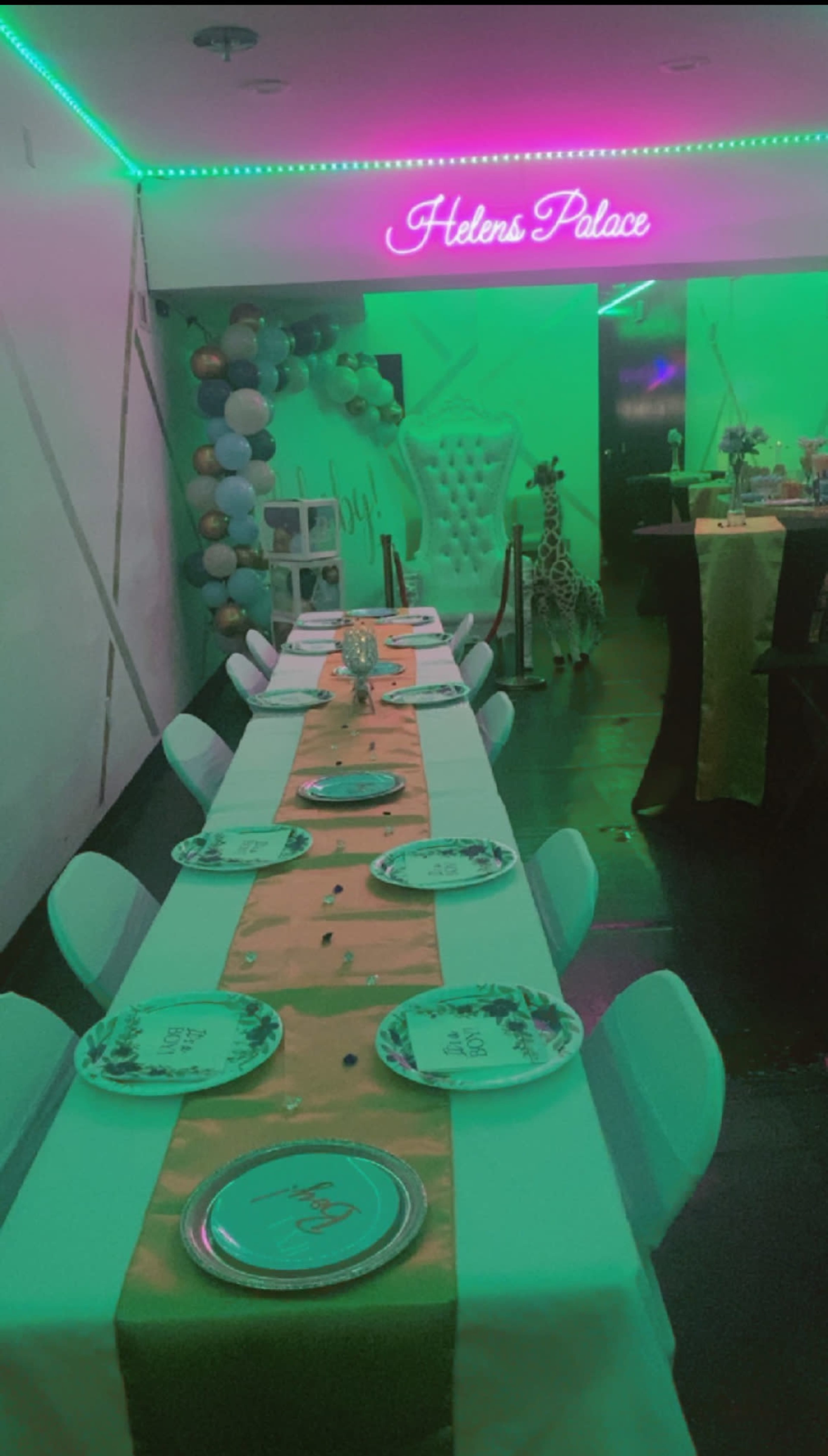 The image shows a decorated dining area with a long table set with plates, a gold table runner, and balloons in the background, illuminated by green lighting.